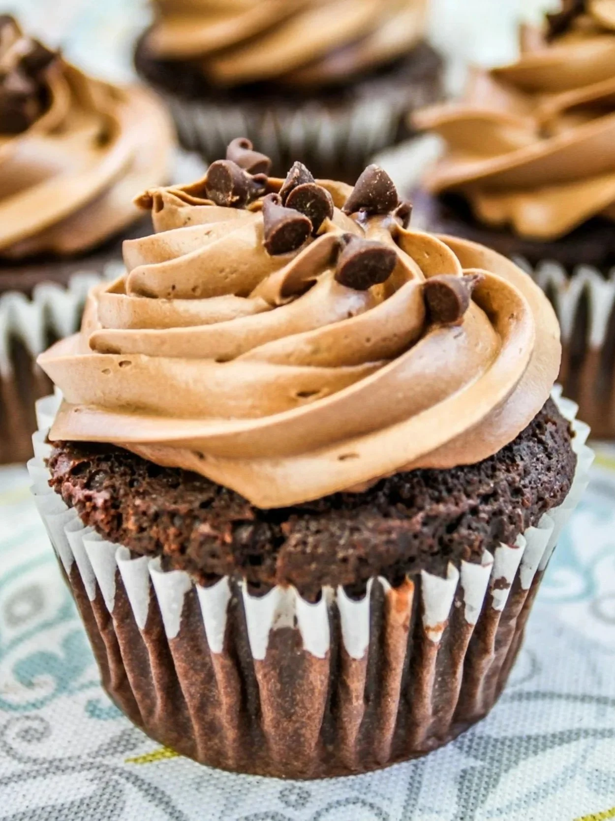 Close-up of a chocolate cupcake with swirled chocolate frosting topped with chocolate chips in a white cupcake liner on a patterned cloth.