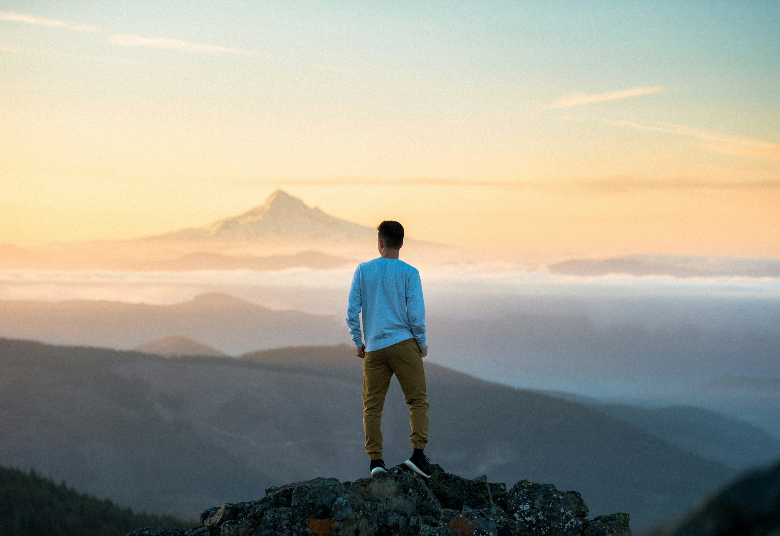 A person standing on a rocky ledge overlooking a foggy landscape with mountains in the distance and a snow-capped volcano