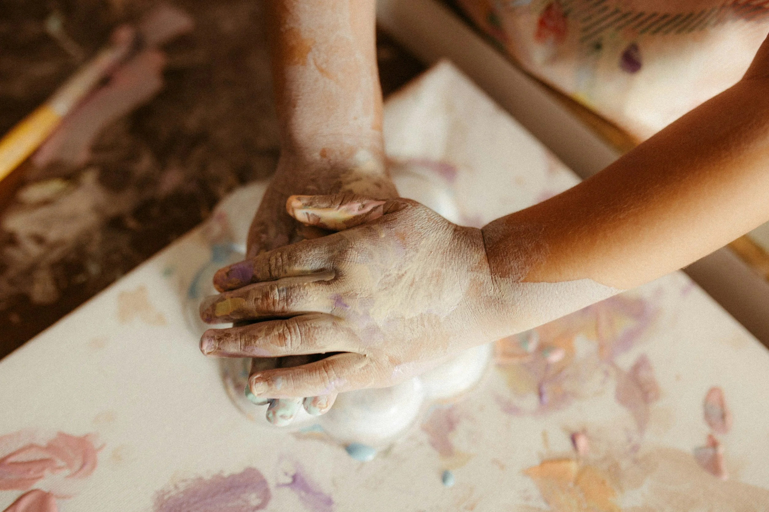 Close-up of two hands covered in multicolored paint, holding each other over a painted surface with splatters of paint.