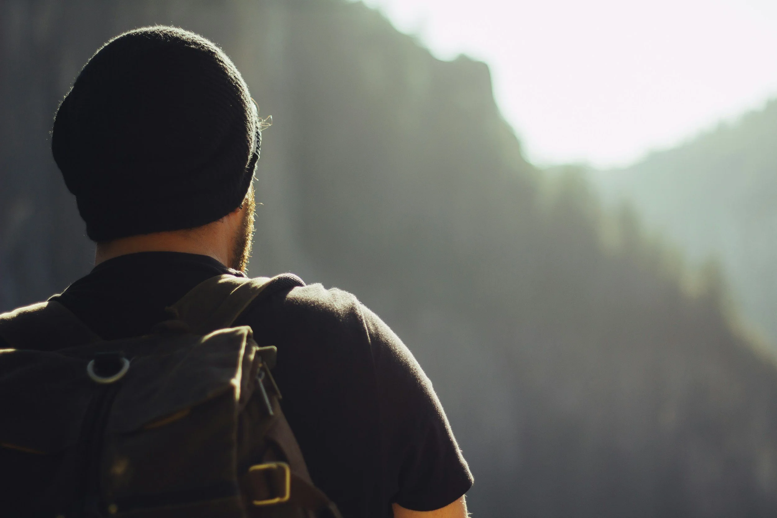 A person wearing a black beanie and glasses, with a backpack, standing outdoors facing a bright light or sunrise.