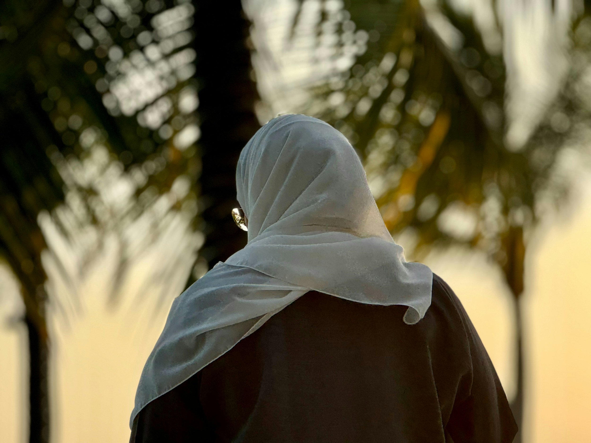 Close-up of a person wearing a white head covering, viewed from behind with blurred green foliage in the background.