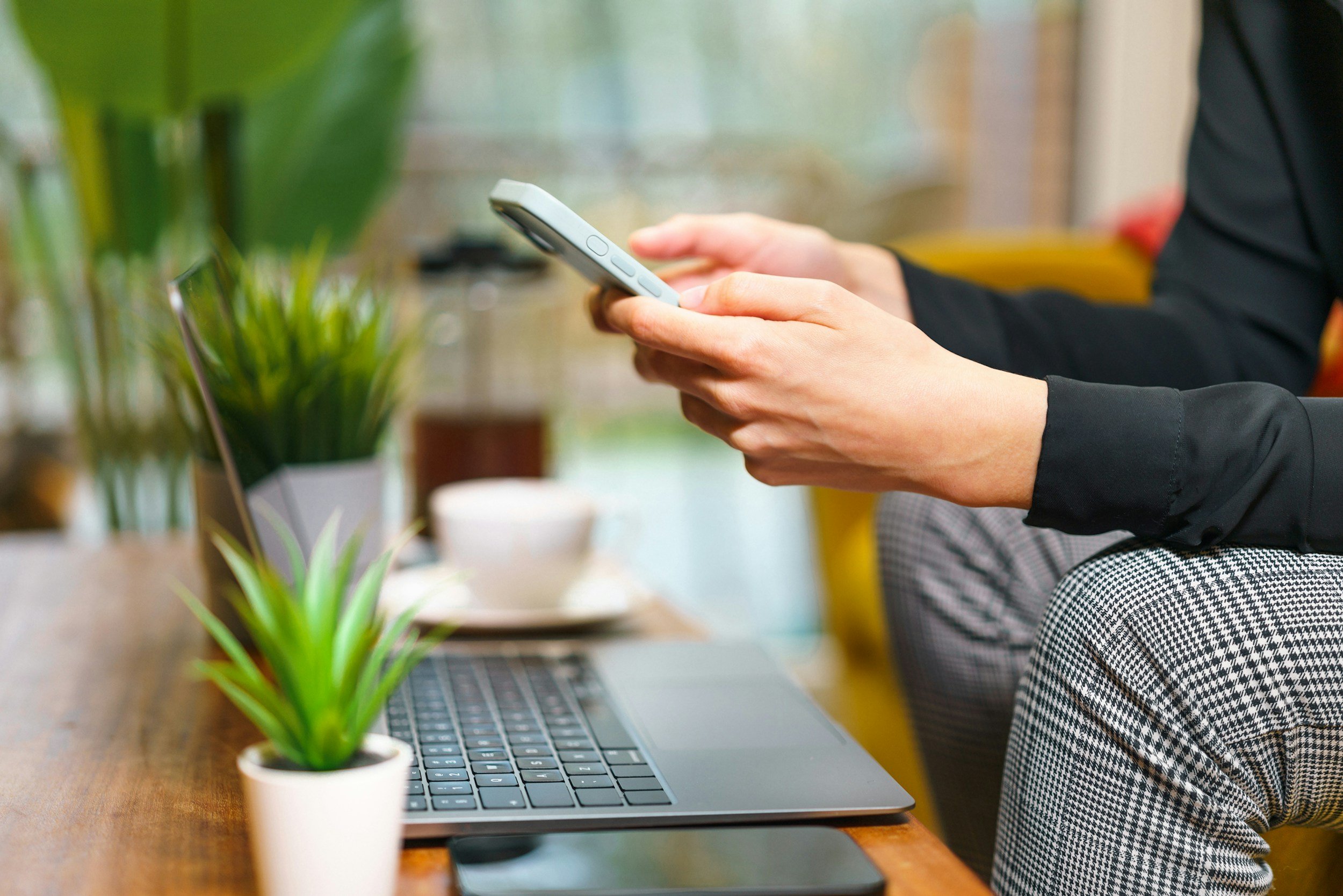 Person using smartphone at a wooden table with a laptop and potted plants.