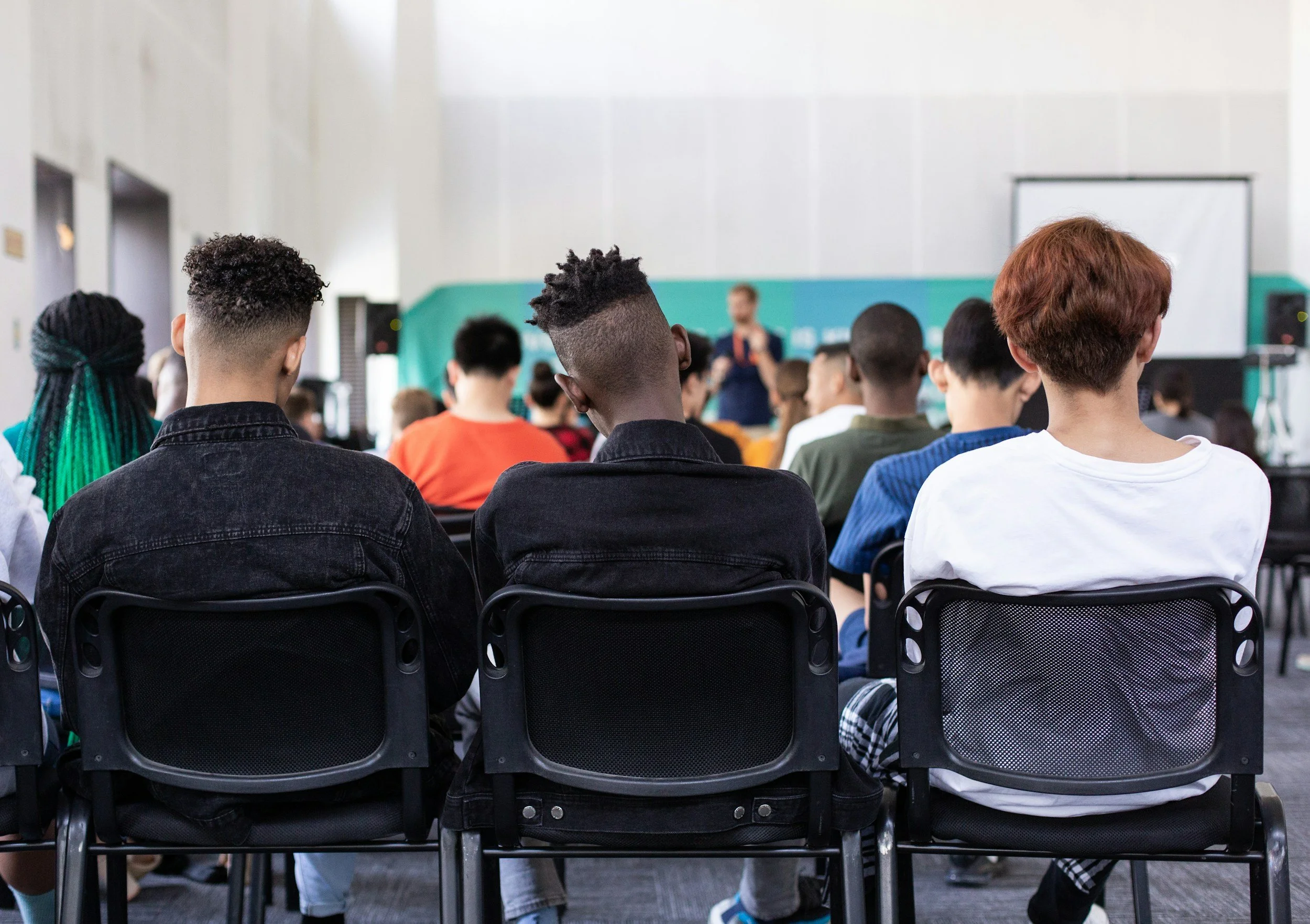 A diverse group of students seated and facing a teacher or speaker in a classroom or lecture hall.