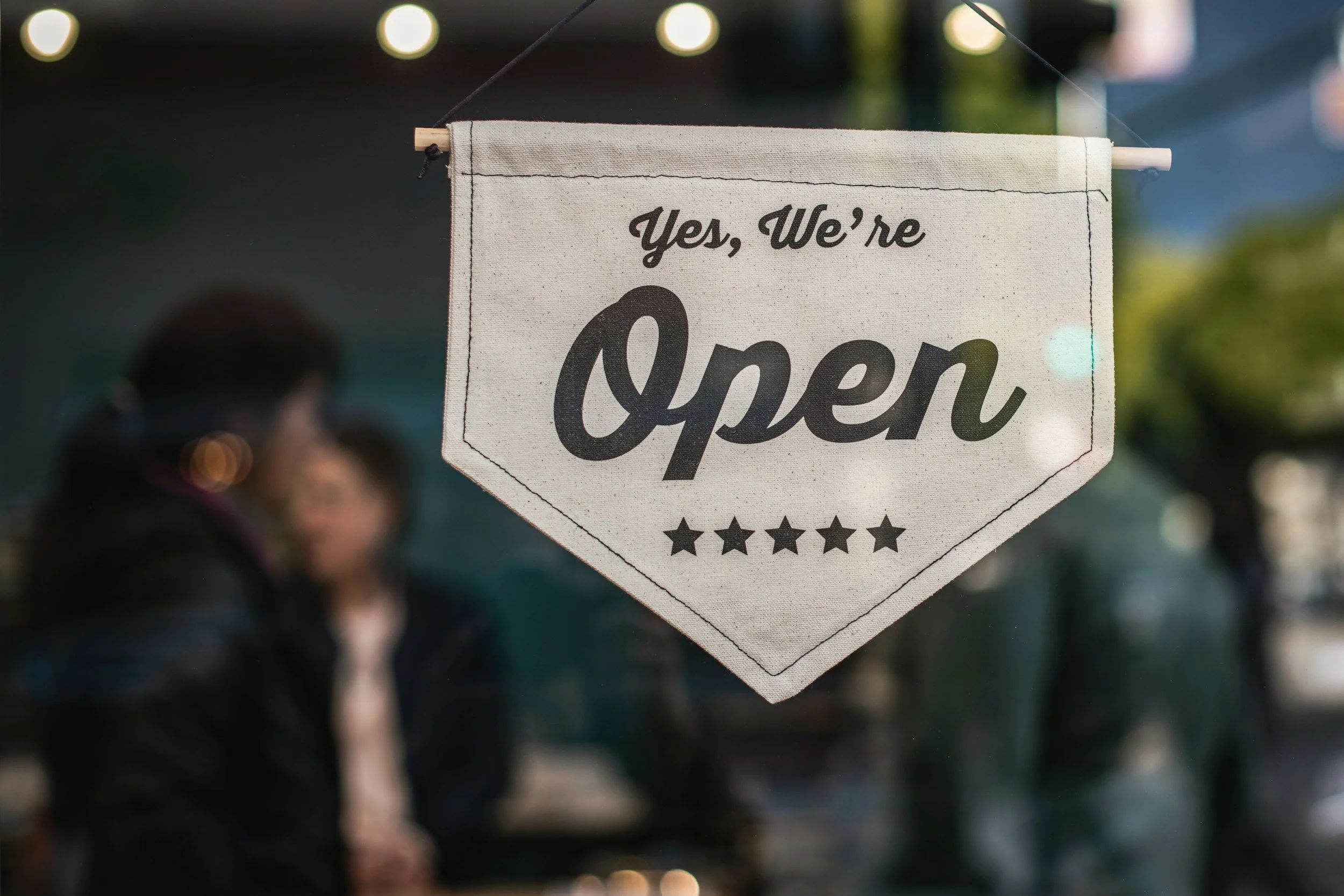 A sign in a store window with text that reads, 'Yes, We're Open' and five stars at the bottom. The background shows blurred people and outdoor lights.