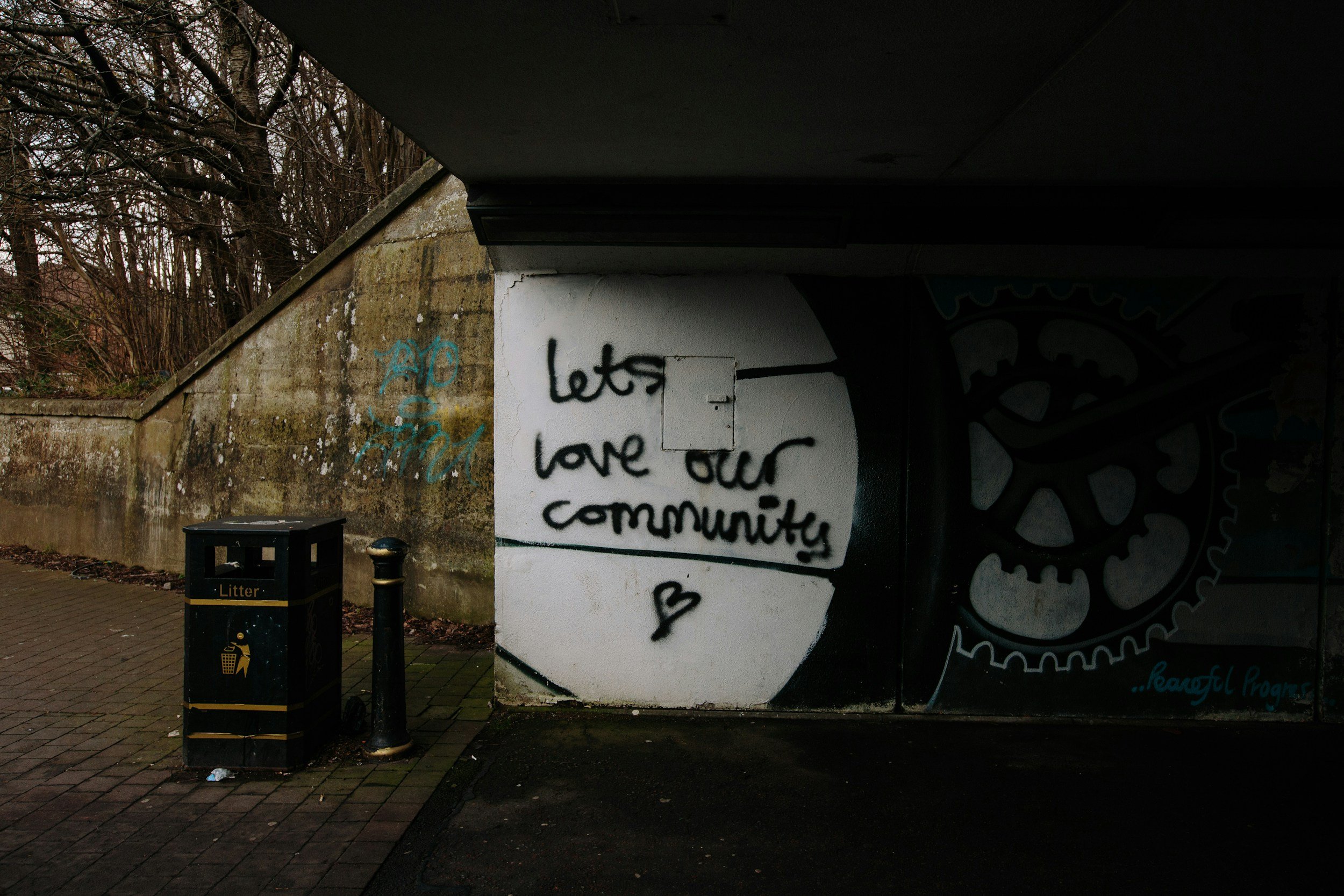Graffiti on a wall under a bridge with the message 'let's love our community.' There is a trash bin labeled 'Litter' nearby, and leafless trees are visible in the background.