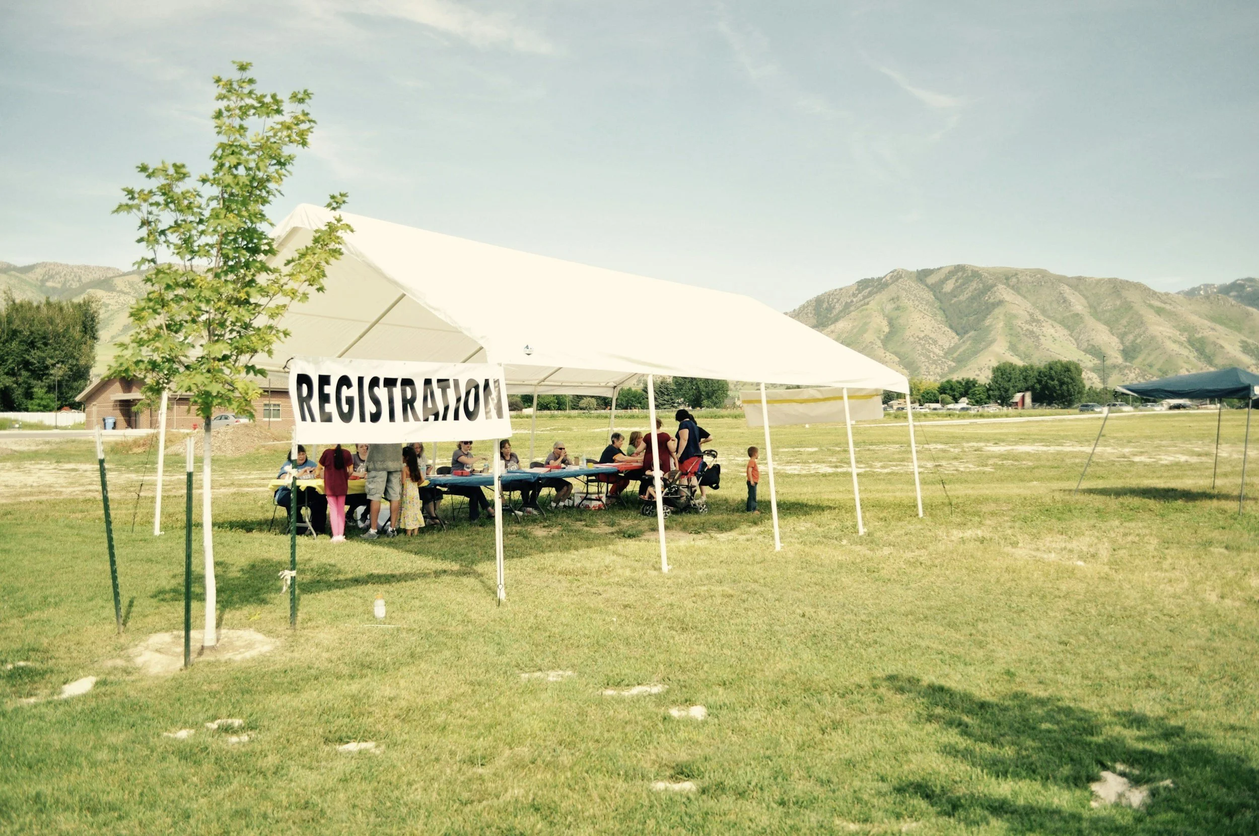A group of people sitting at a registration table under a white canopy tent in a field with green grass, with mountains and a blue sky in the background.