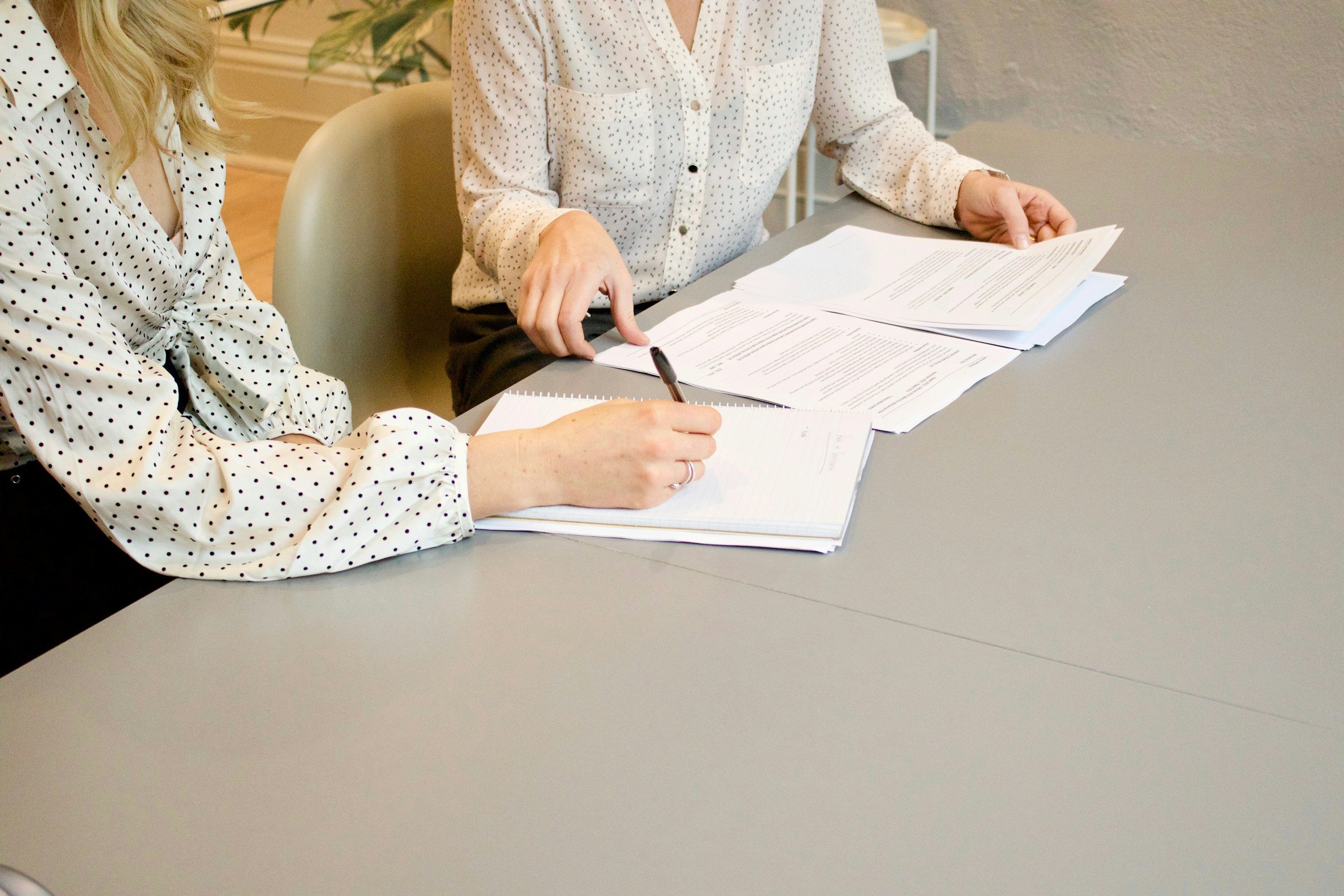 Two women sitting at a table, reviewing and taking notes on documents, in an office setting.
