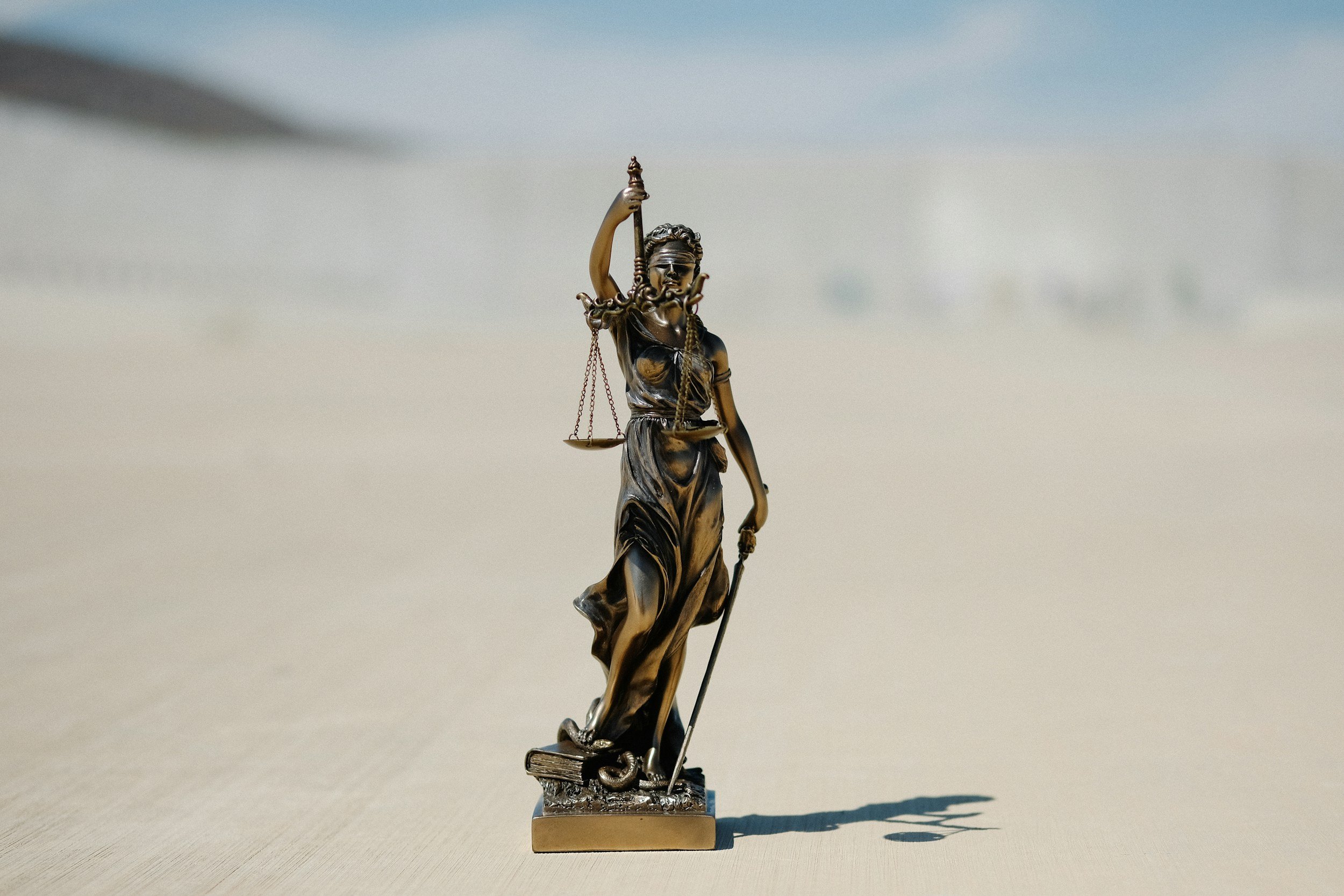 A small metal sculpture of Lady Justice holding scales in her left hand and a sword in her right hand, standing on a rectangular base with a book, cast a shadow on a light-colored surface.