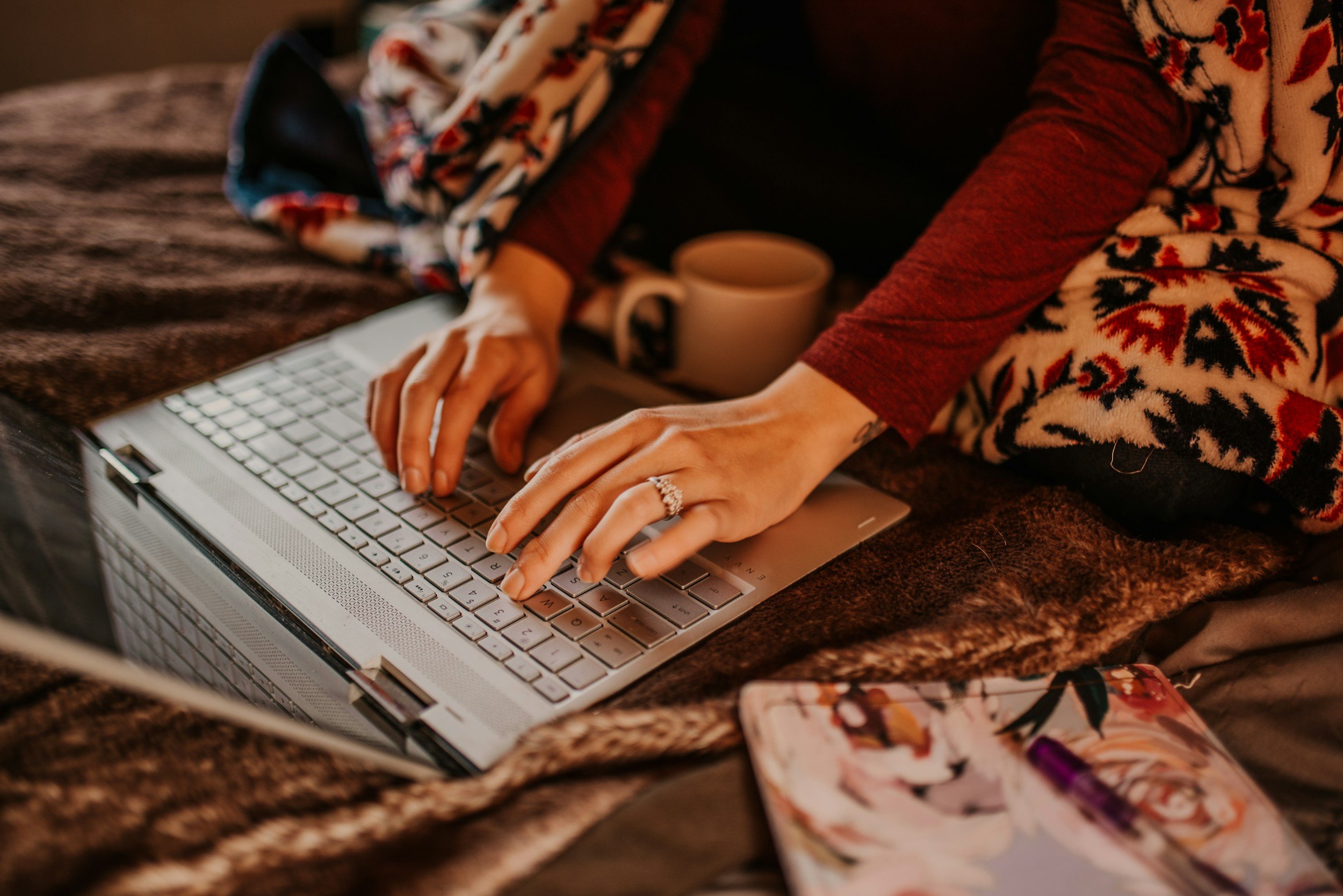 Person typing on a laptop keyboard with a coffee mug nearby, surrounded by floral-patterned items.