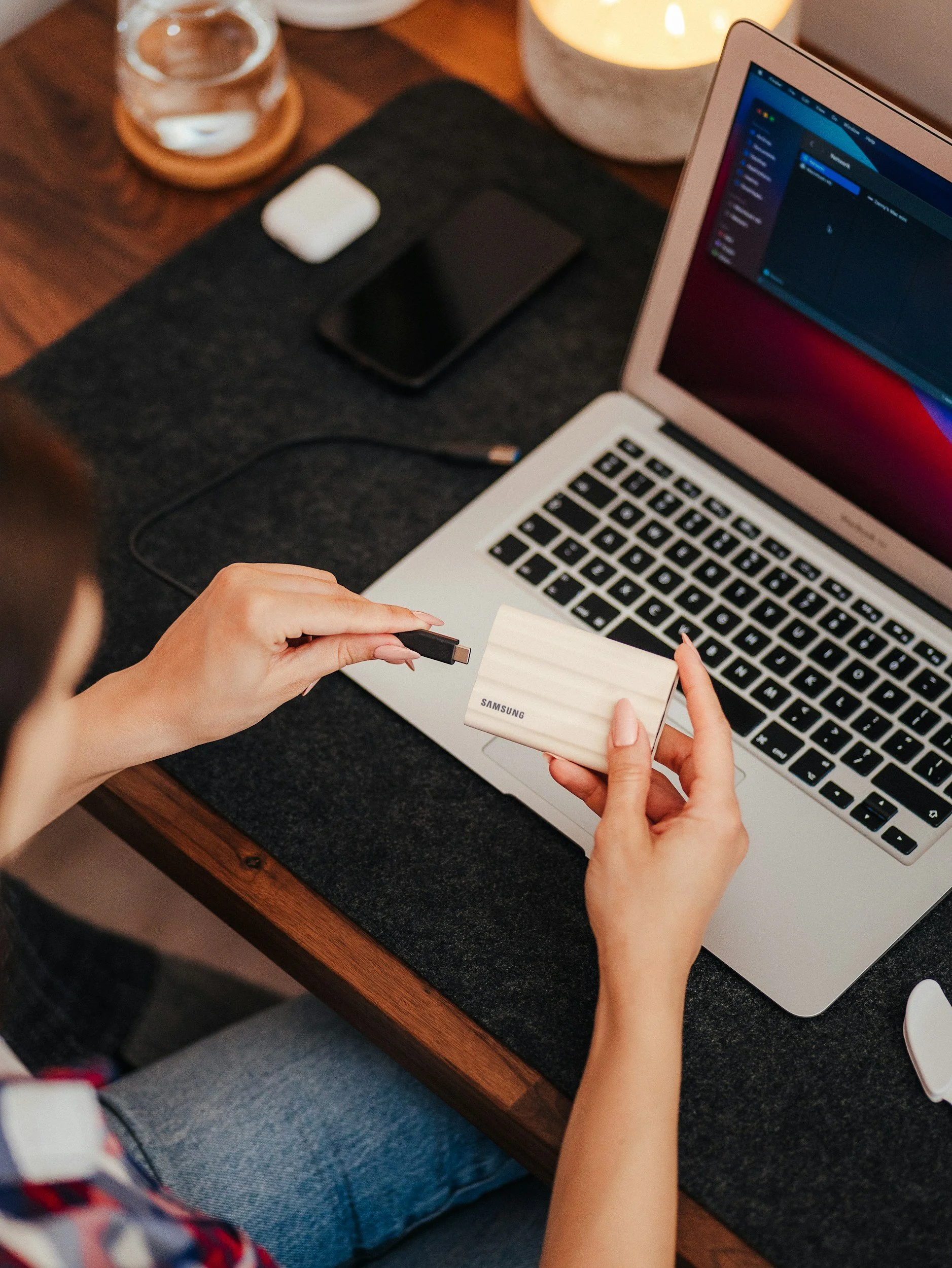 A person is holding a Samsung USB flash drive and a Samsung external hard drive over a laptop keyboard, on a wooden desk with a glass of water on a coaster, a lamp, and a closed black notebook nearby.