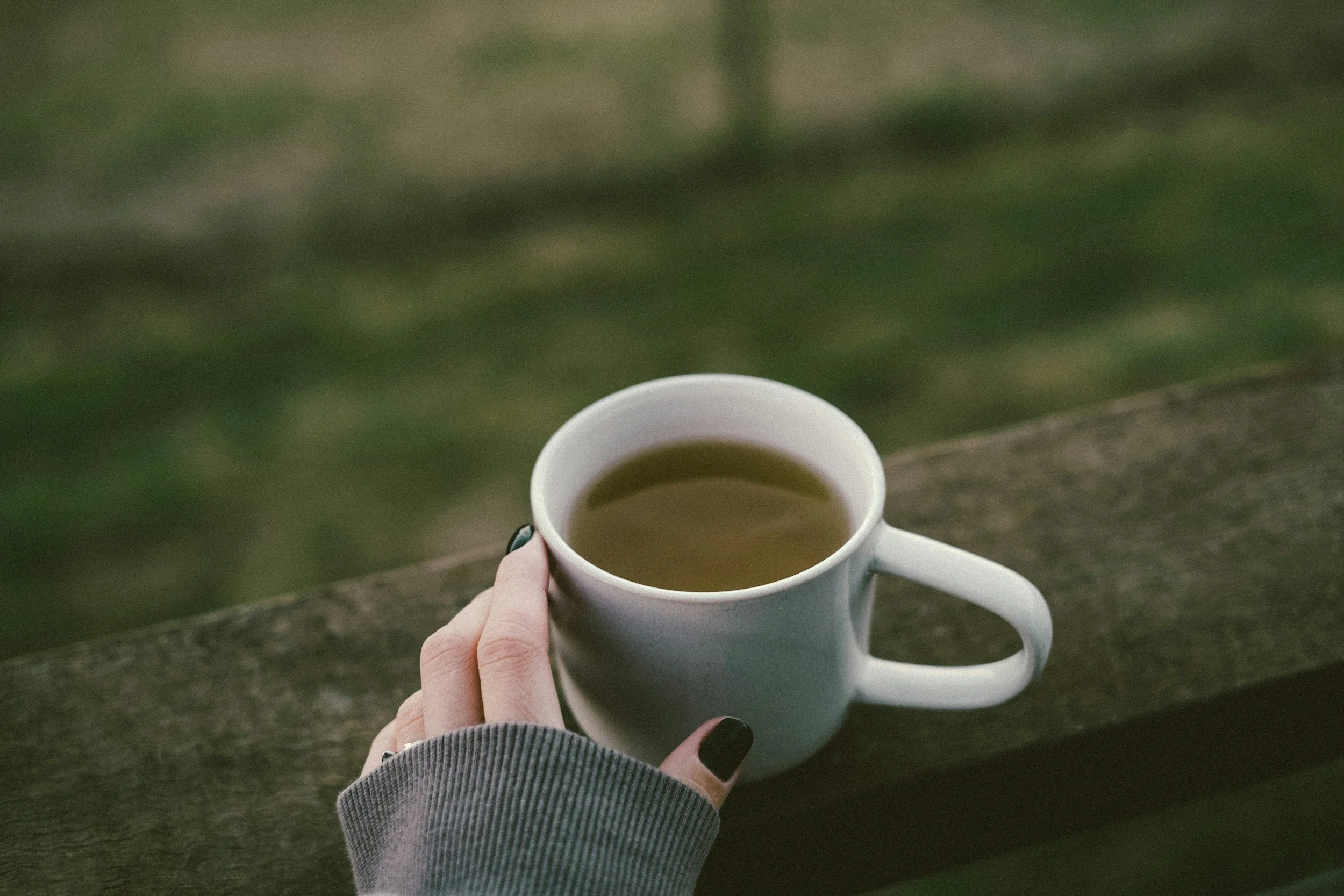 A person holding a white mug filled with a hot beverage outdoors, with a blurred green background and part of their hand and sleeve visible.