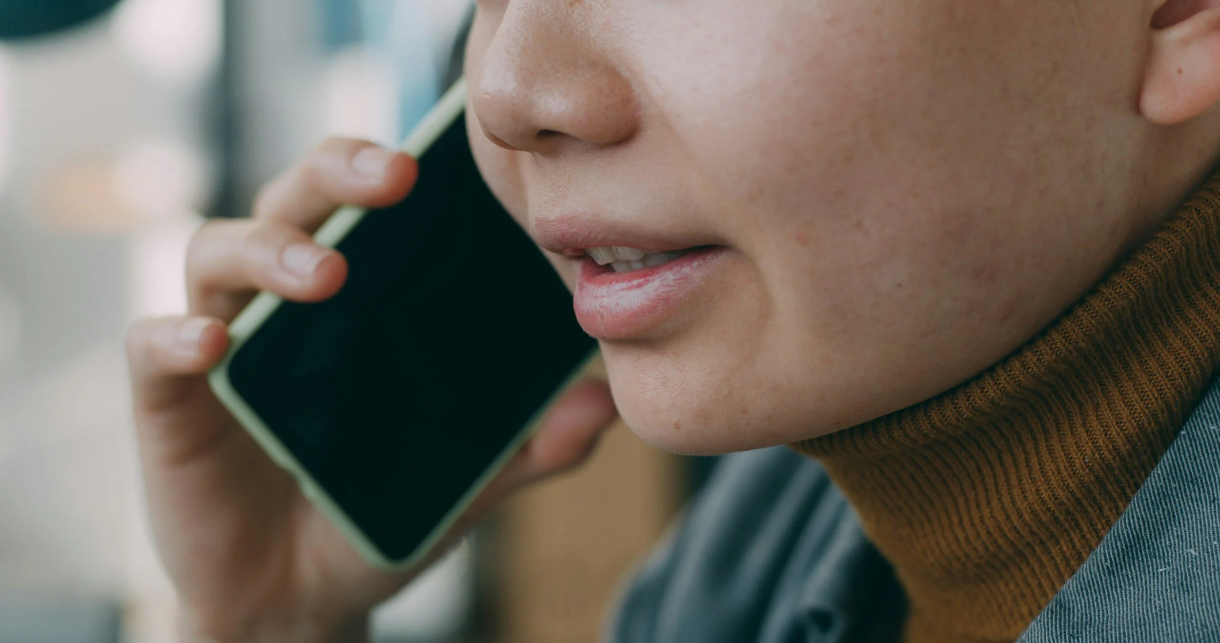 Close-up of a person holding a phone to their ear, smiling, with part of their face visible, wearing a brown scarf and striped shirt.