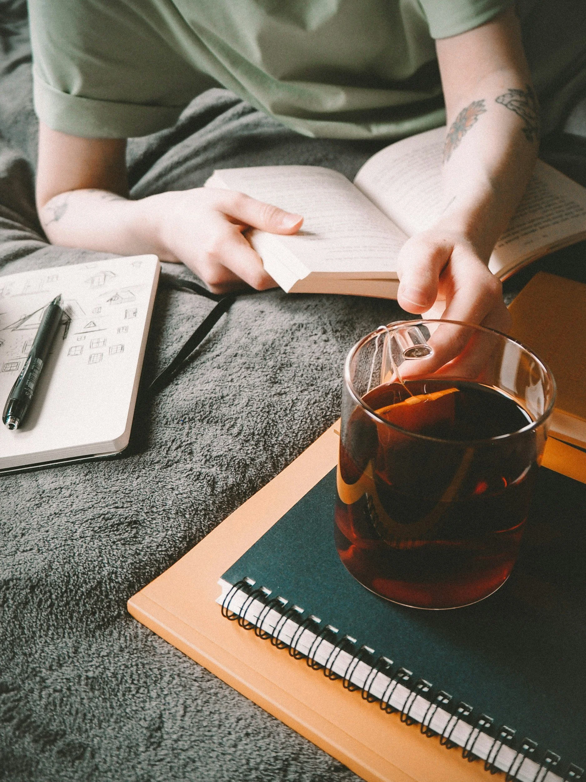 A person sitting on a gray carpet, reading a book, with a glass of dark beverage, notebooks, and sketches on the surface in front of them.