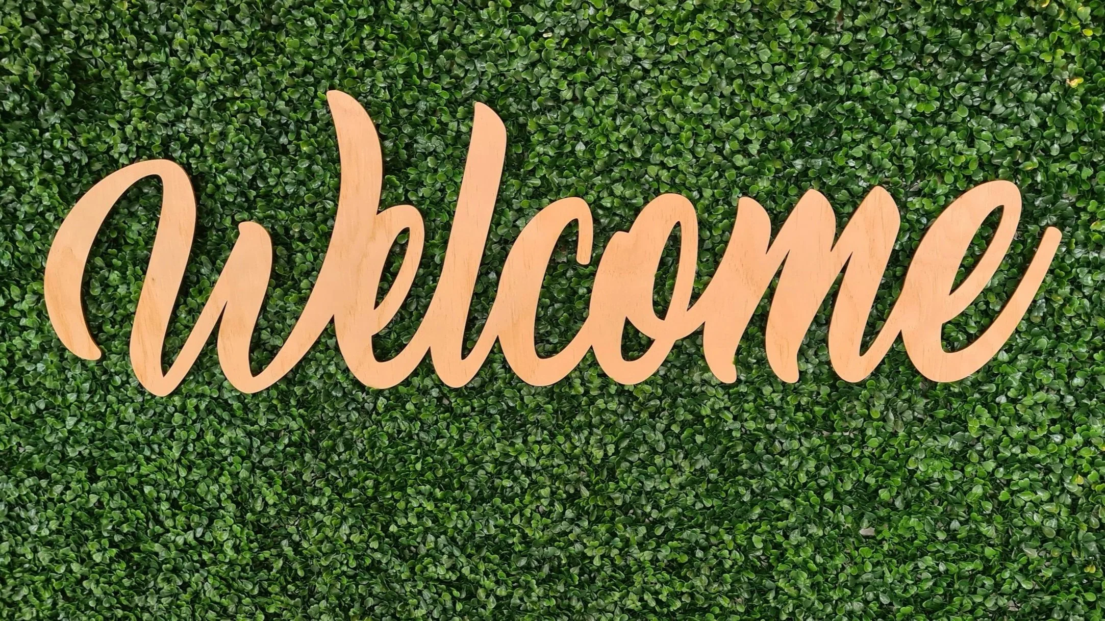 Wooden sign with the word 'welcome' in cursive font, mounted on a lush green hedge background.