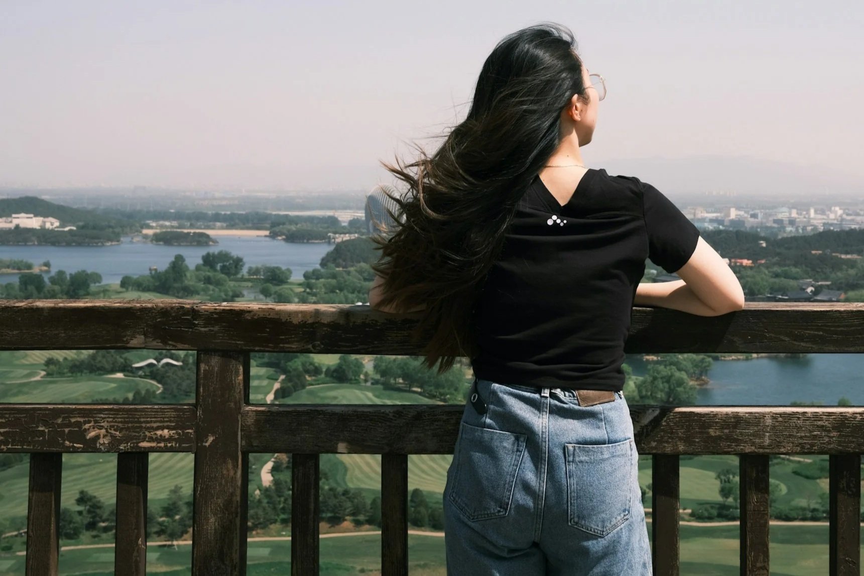 A woman with long dark hair wearing glasses, a black t-shirt, and blue jeans is standing on a viewing platform, looking out over a landscape with a river, trees, and a city in the distance.