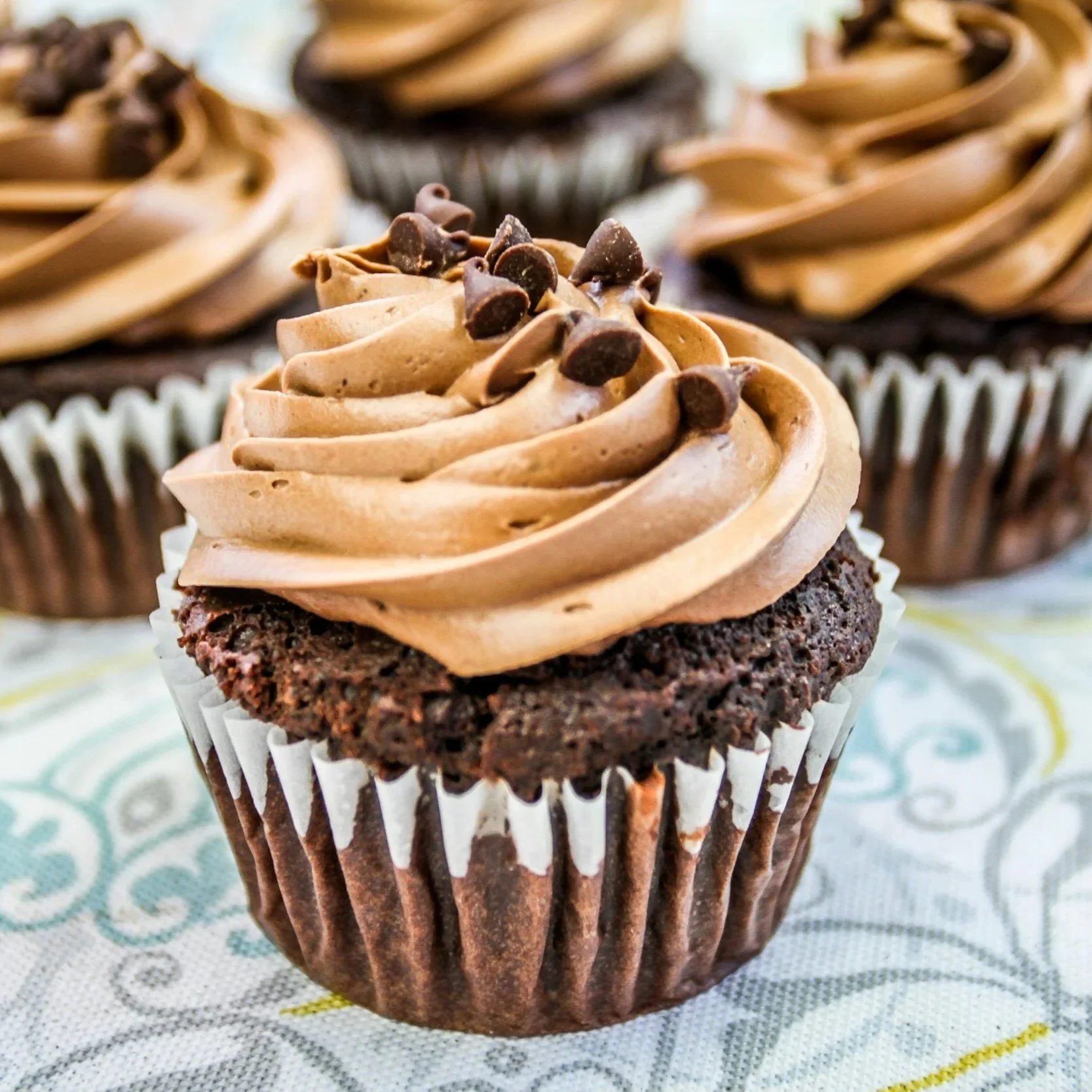 Chocolate cupcake with light brown frosting and chocolate chips on top, placed in a white cupcake liner with a black paper wrapper, on a patterned cloth background.