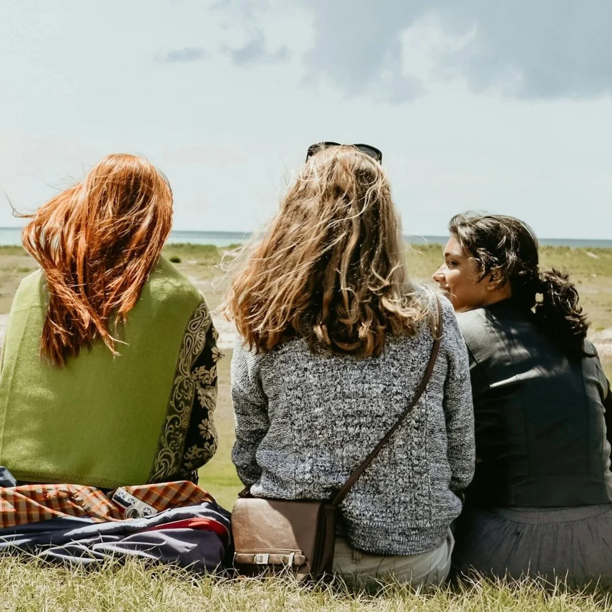 Three women sitting on grass near the coast, looking out at the ocean on a partly cloudy day.