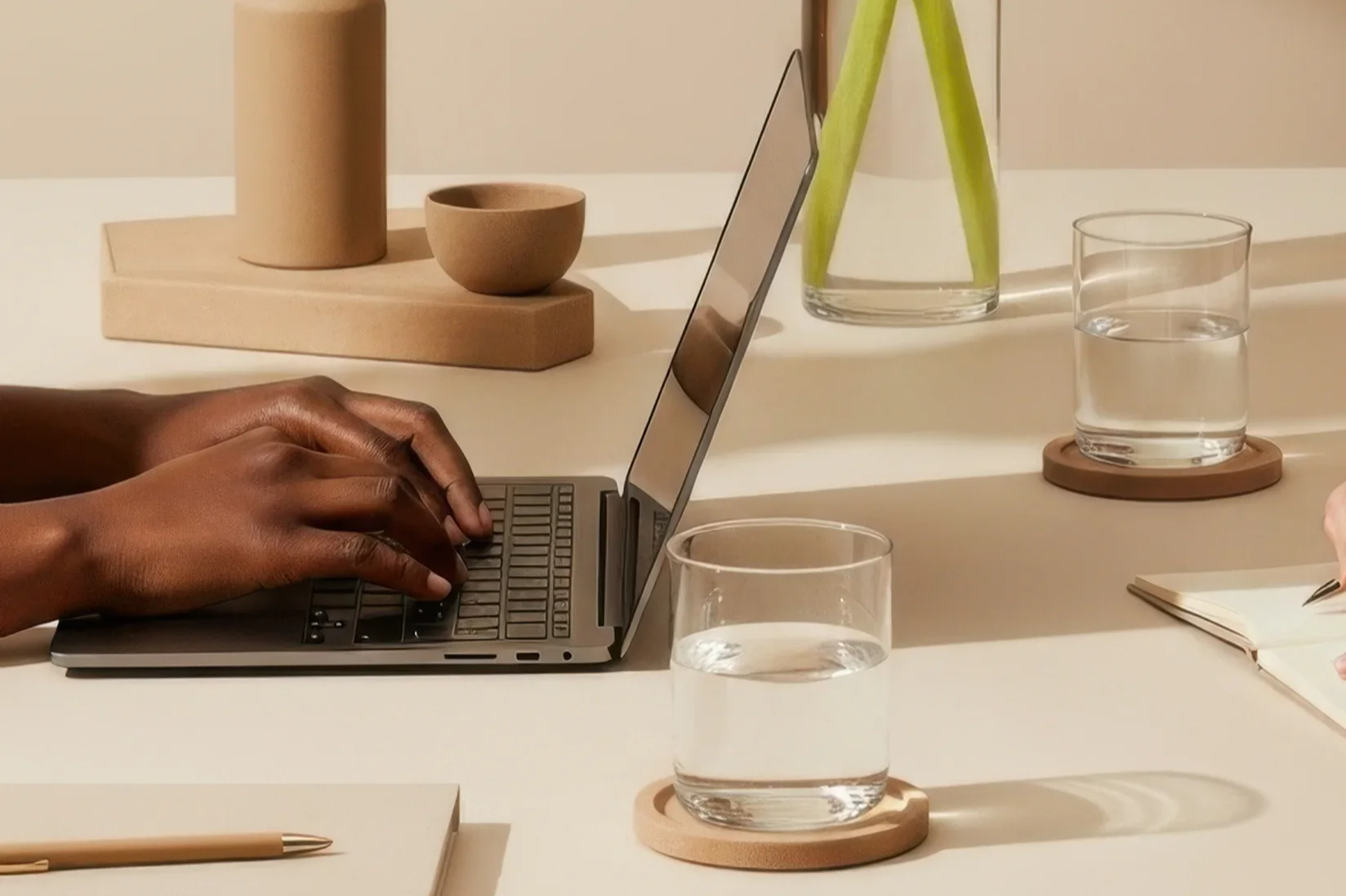 Person typing on a laptop on a beige desk with two glasses of water, a notebook, a pen, a vase with green stems, and decorative objects in earthy tones.