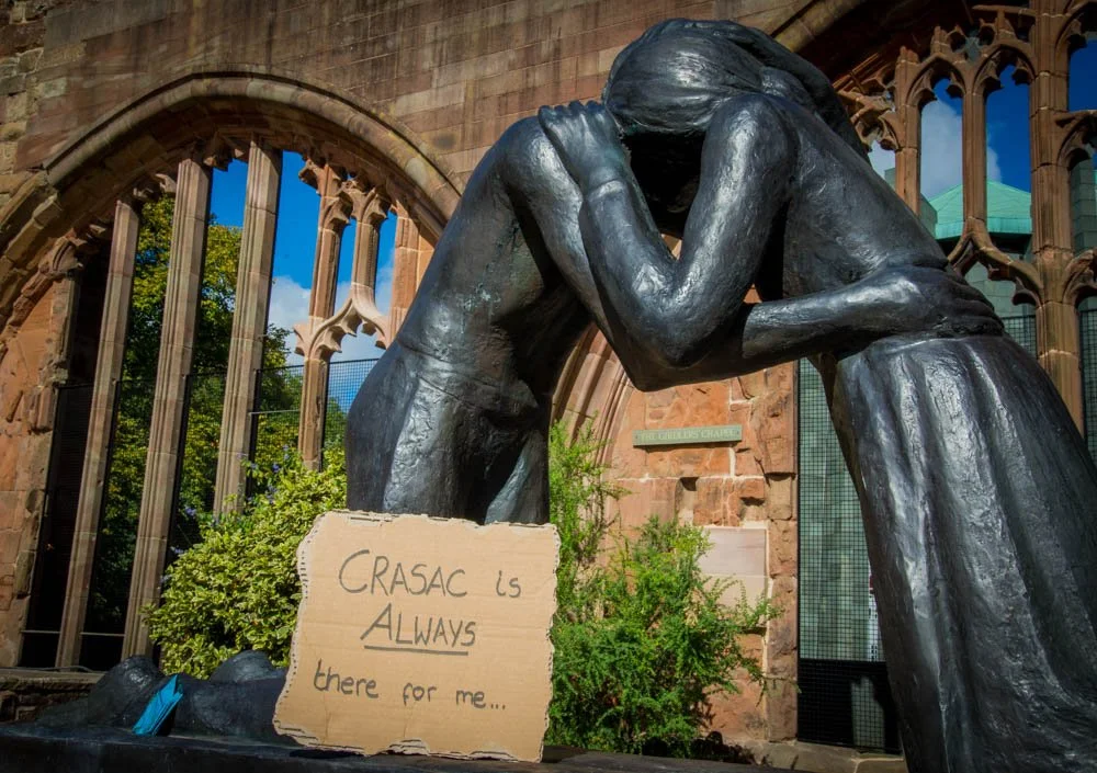 A bronze sculpture of two people embracing, with a sign that reads, "CRASAC is ALWAYS there for me..." in front of a brick building with large arched windows and green trees in the background.