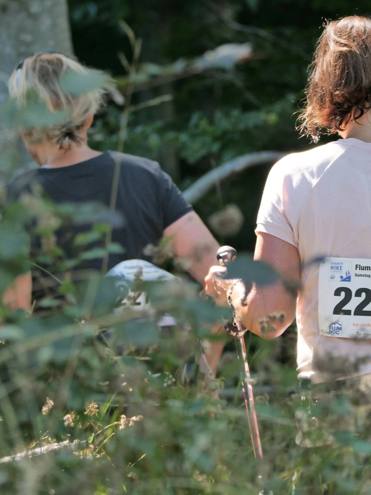 Two people participating in a charity hike through a forested area, wearing race bibs with numbers and standing among greenery.