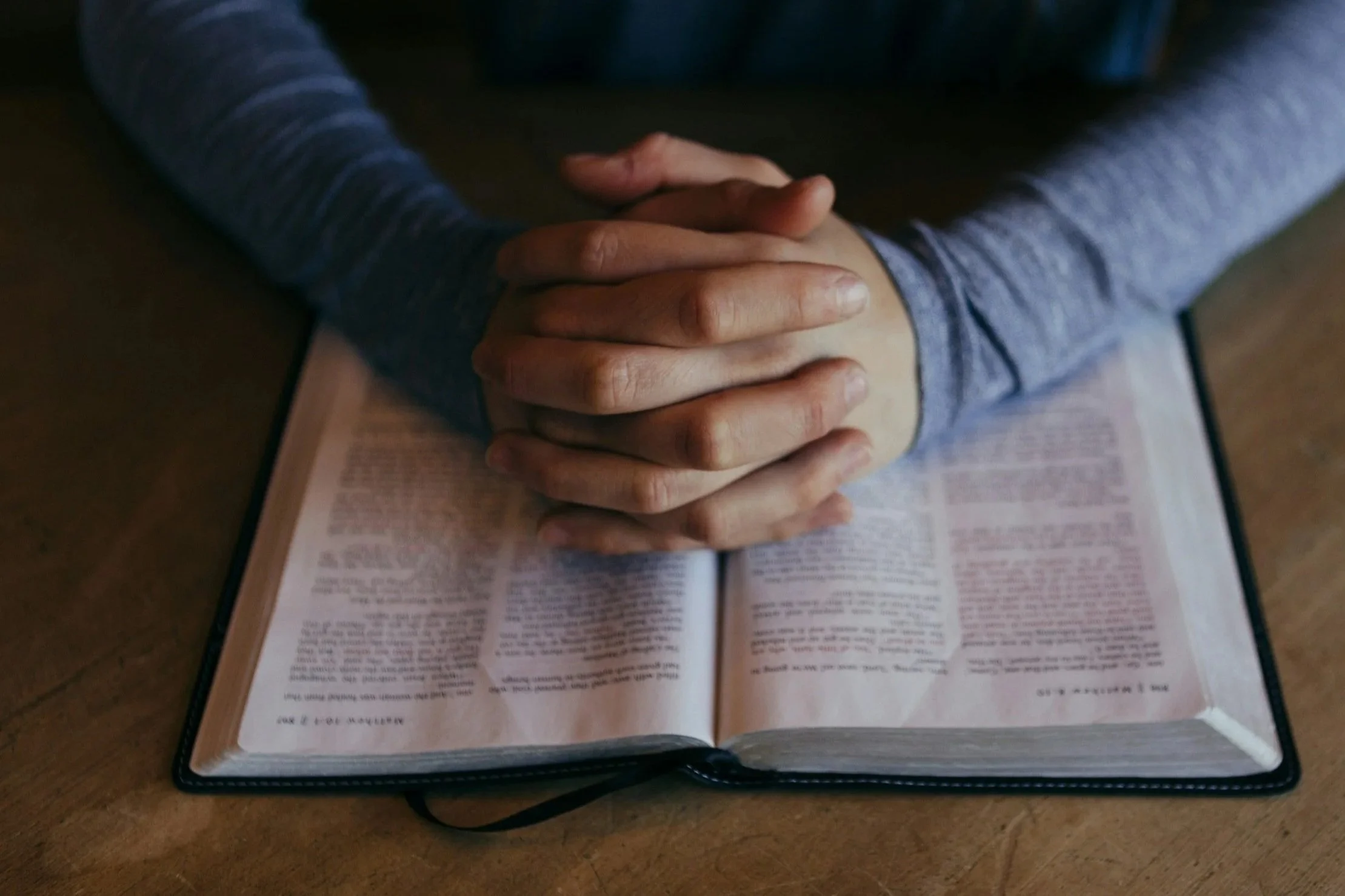 Person with clasped hands resting on open Bible or religious book on table.