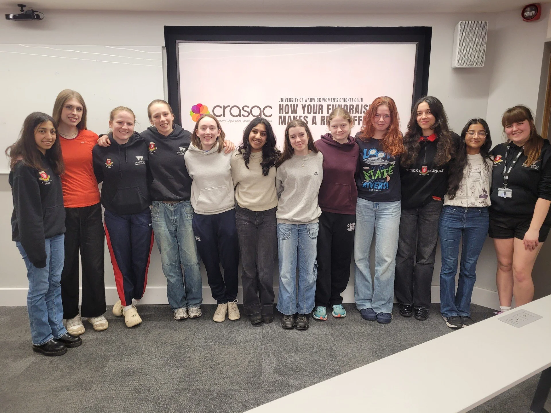 A group of girls standing in a classroom in front of a presentation screen that reads "HOW YOUR FUNDRAISERS MAKES A DIFFERENCE," with a logo for CRSAC and university of Warwick women's cricket club.