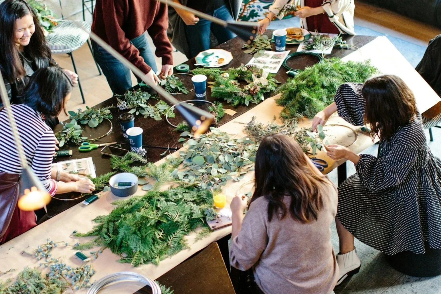 Group of people making Christmas or holiday wreaths with greenery, pine branches, and decorative elements on a large table in a well-lit room.