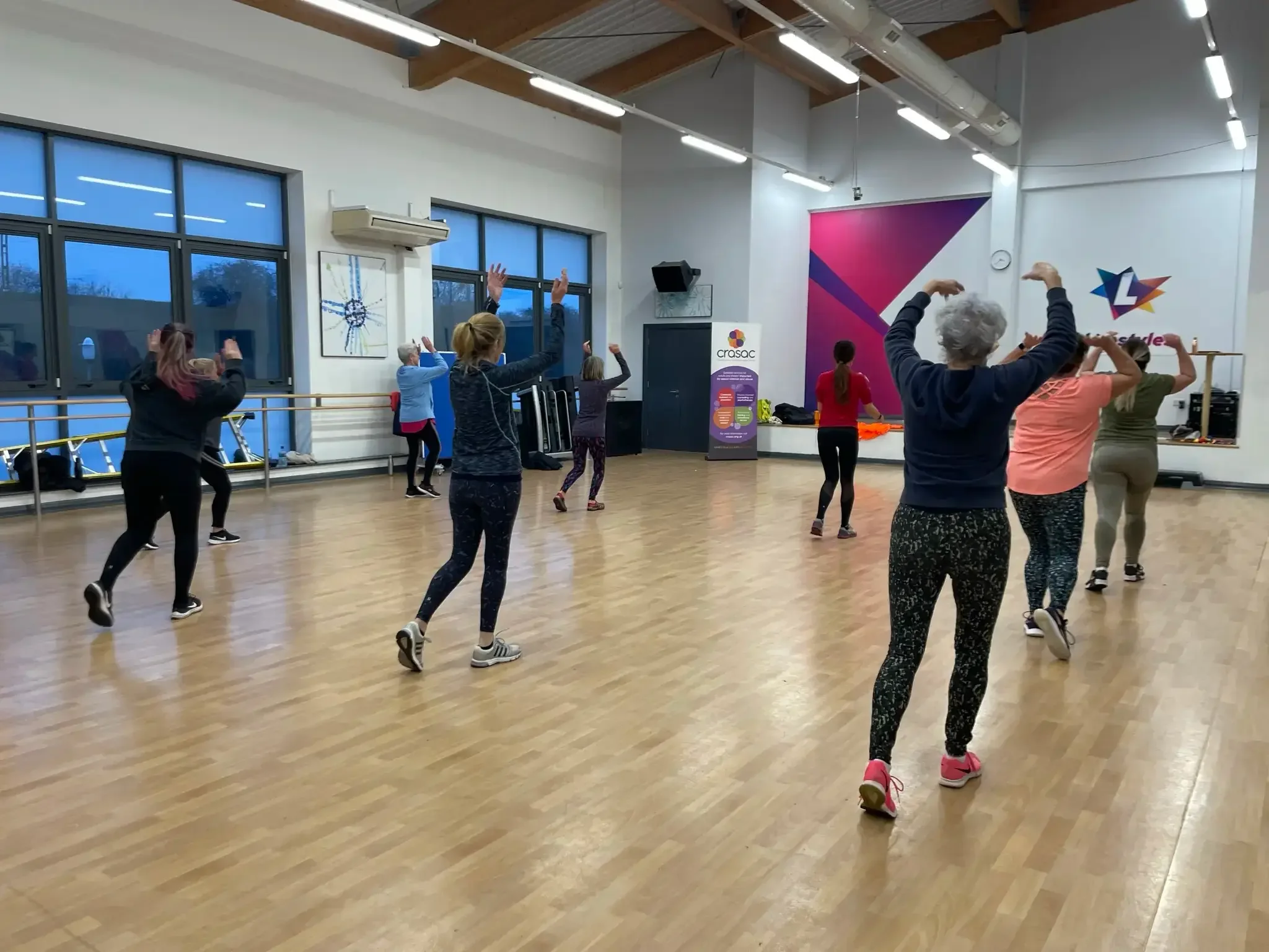 Group of people participating in a fitness class, following instructor in a dance or aerobics studio with large windows, wooden floor, and colorful wall decorations.