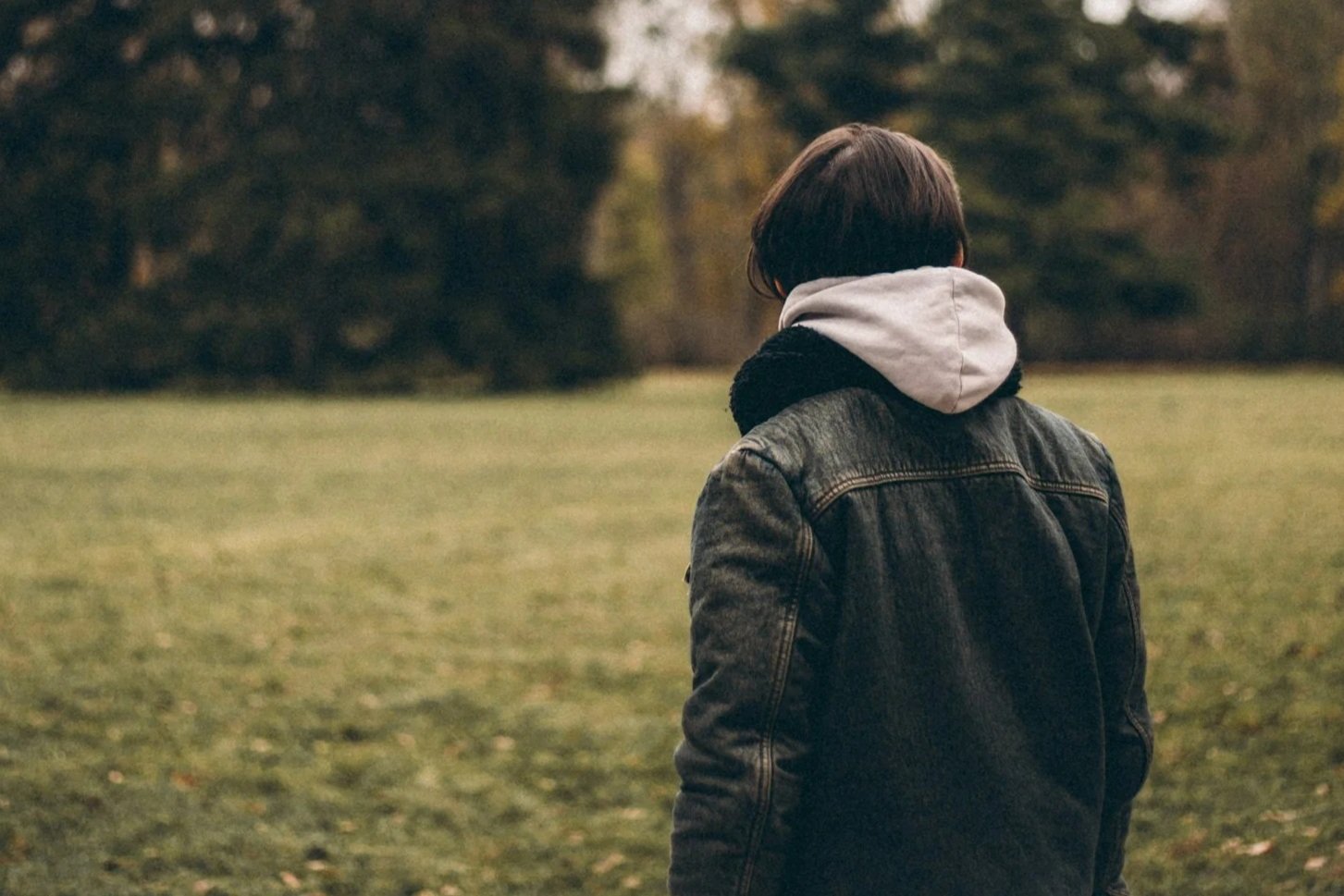 A person with short dark hair standing outdoors in a grassy field, wearing a denim jacket and a hoodie, facing away from the camera, with trees in the background during autumn.