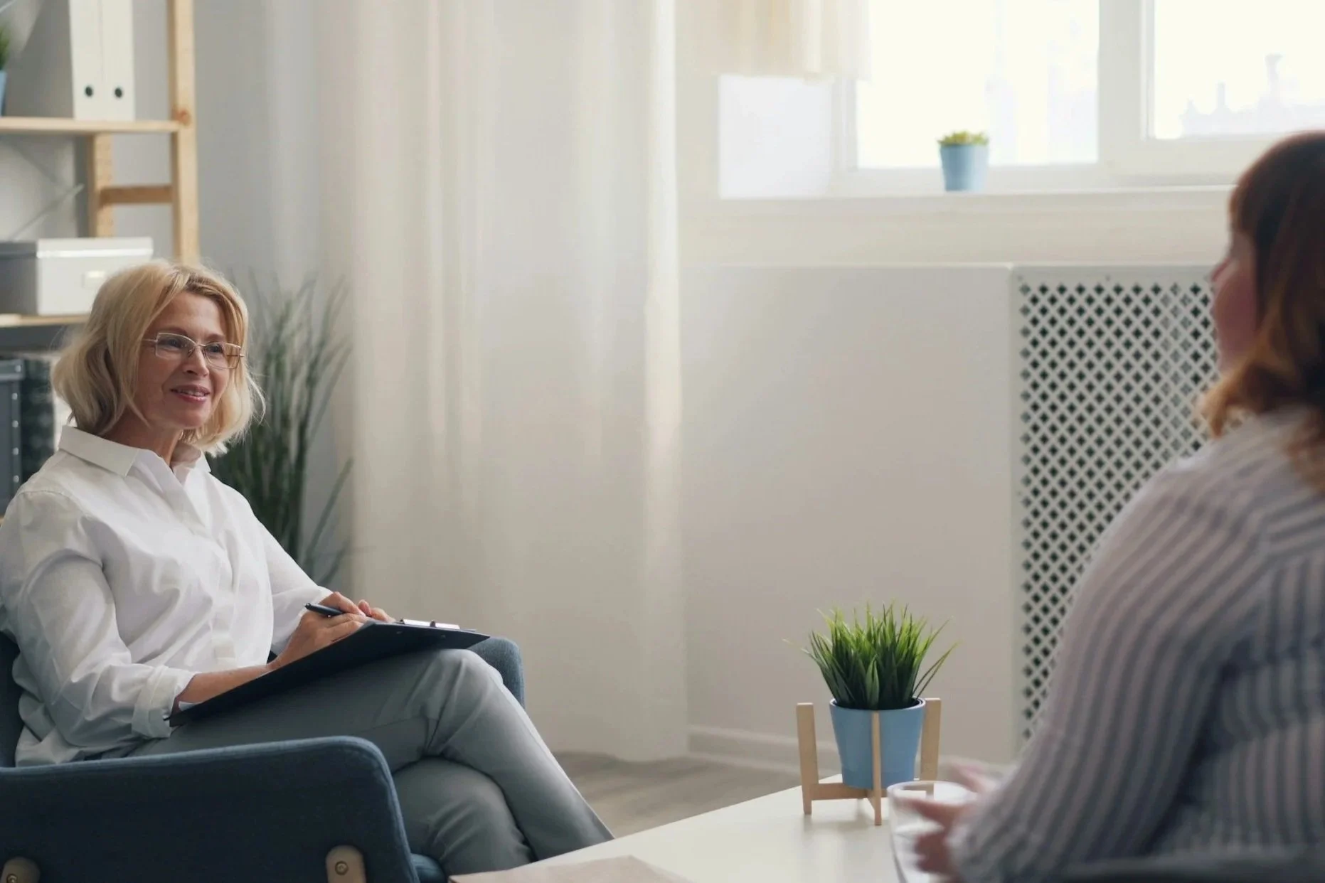 A woman with blonde hair and glasses wearing a white shirt is sitting in a chair, smiling, and holding a clipboard with papers. She is in a bright room with white walls, near a window, with a small potted plant on the table in front of her. Another woman is partially visible, sitting across from her.