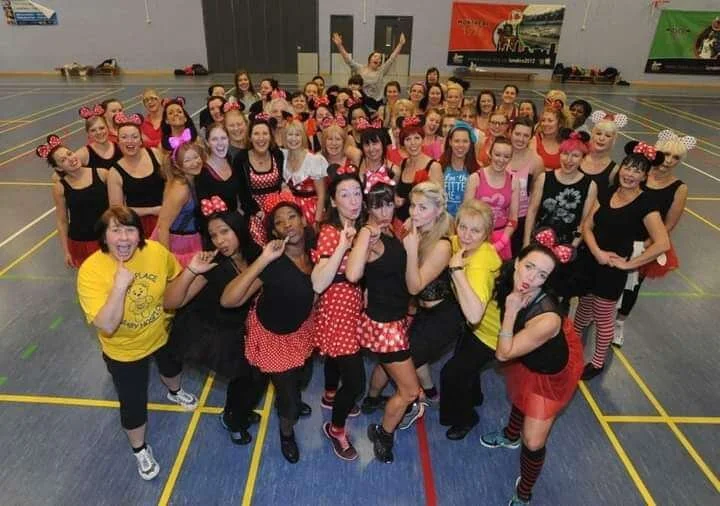 Group of women and girls dressed in Minnie Mouse-themed costumes, posing in a gymnasium.