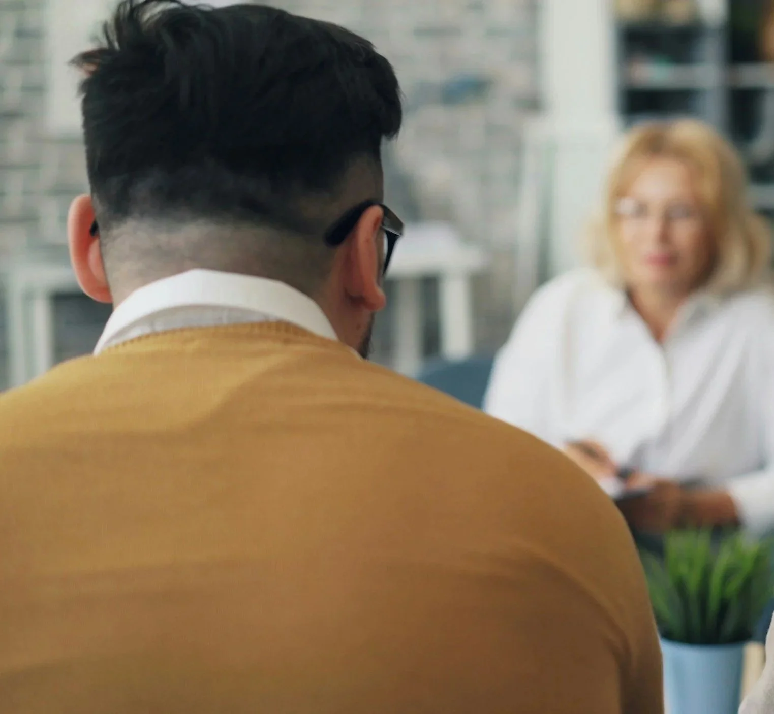 A man with dark hair in a fade haircut wearing glasses and a mustard-colored blazer is having a conversation with a woman with blonde hair and glasses in an office setting.