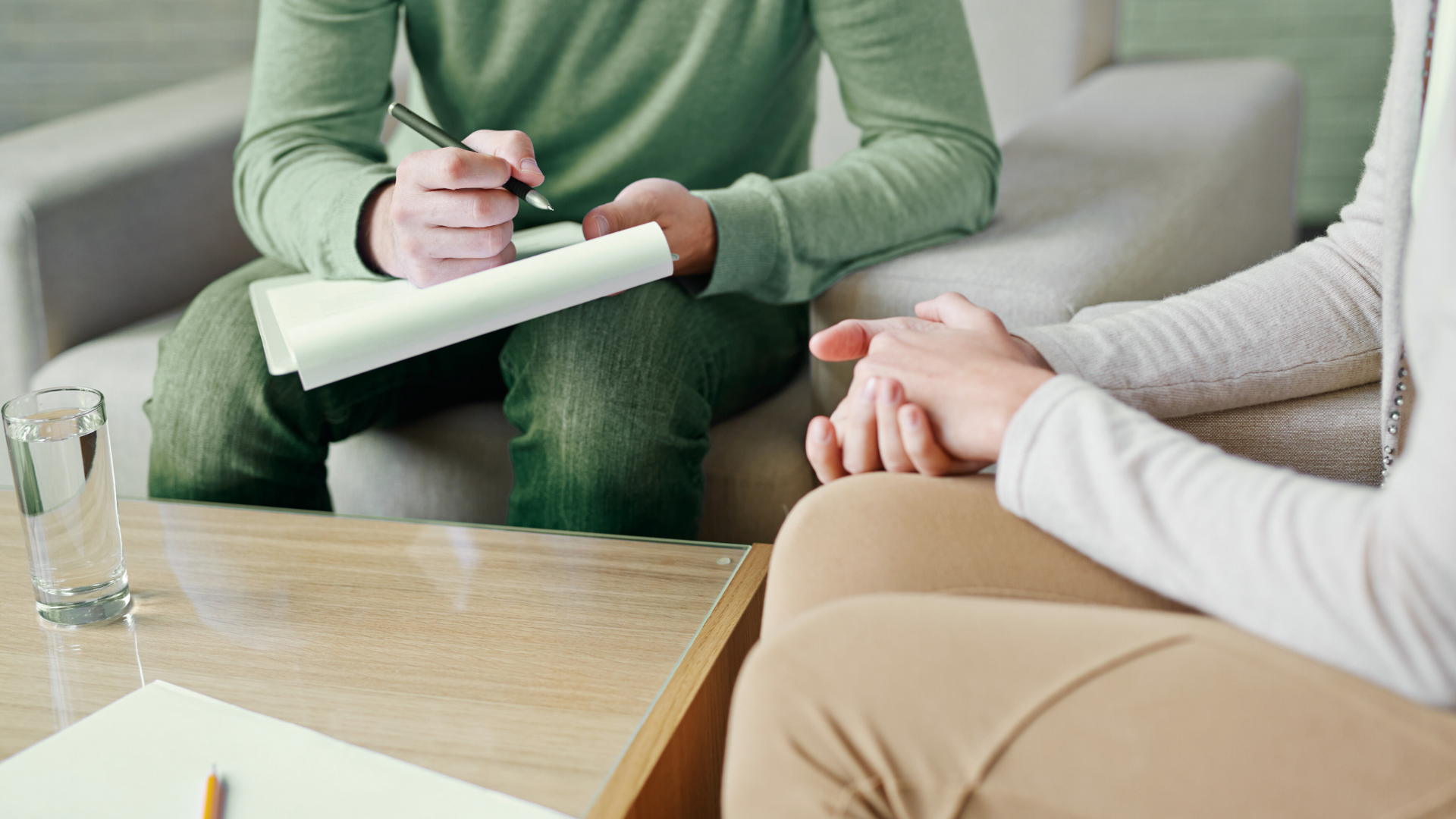 A therapist and a patient sitting on a sofa in a therapy session. The therapist is taking notes on a notepad. A glass of water and some papers are on the table.
