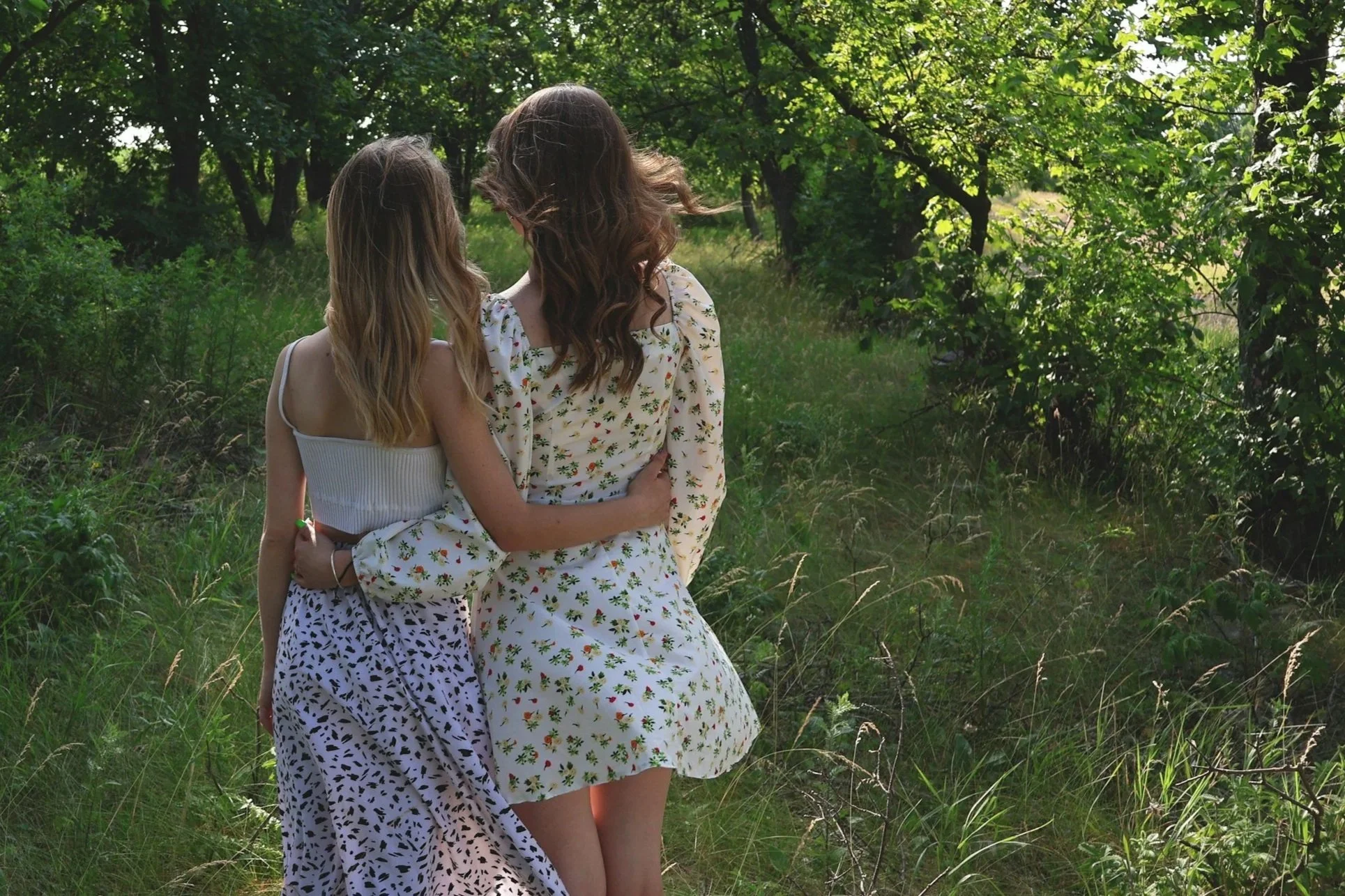 Two women hugging in a green outdoor area with trees and grass, wearing patterned dresses.