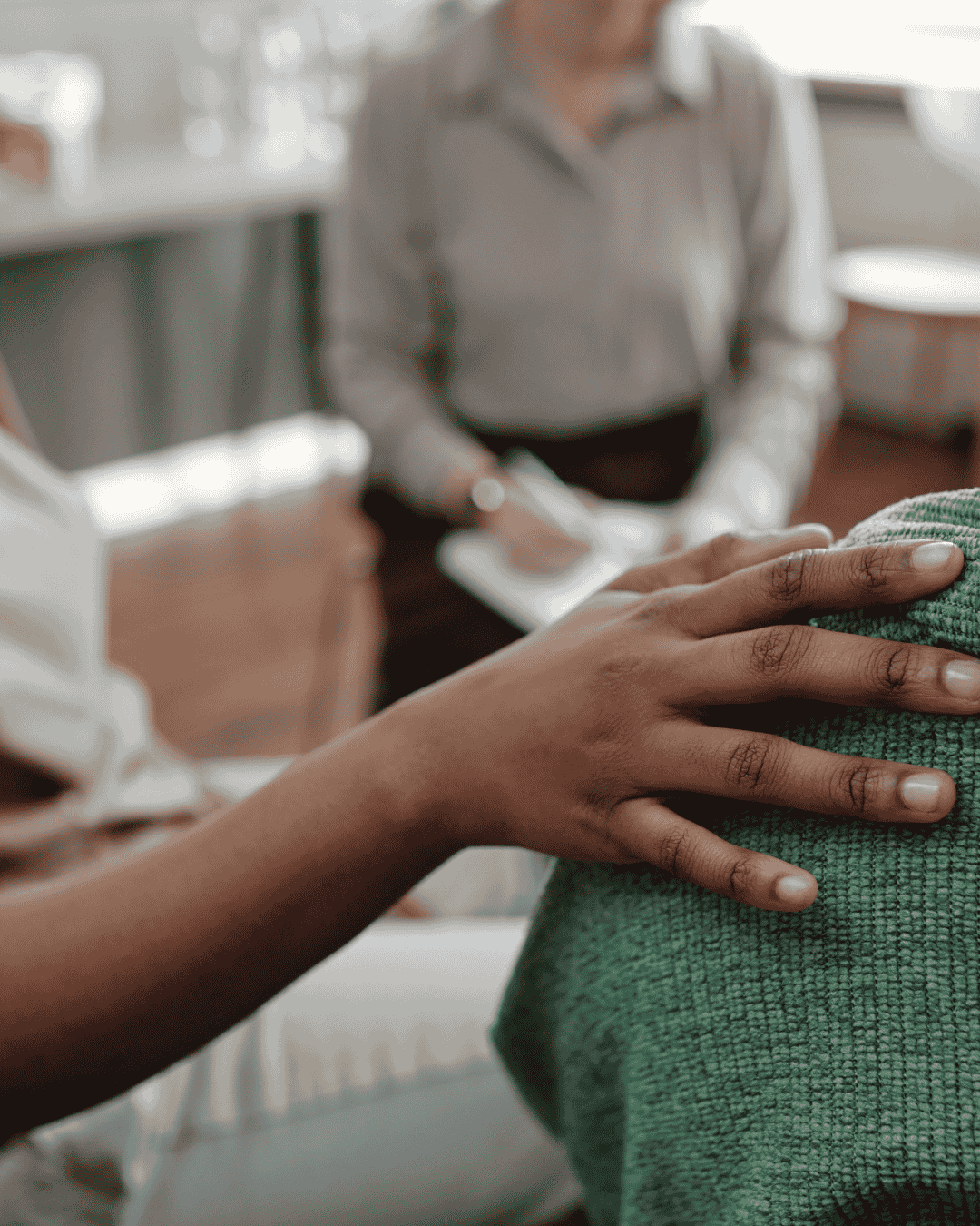 A person touching the shoulder of another person wearing a green sweater in an indoor setting, with a third individual in the background.