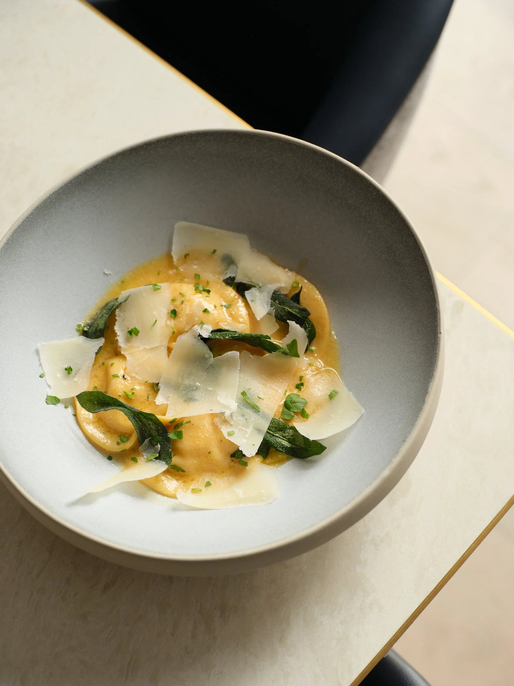 A bowl of ravioli pasta garnished with shaved cheese, and chopped herbs.