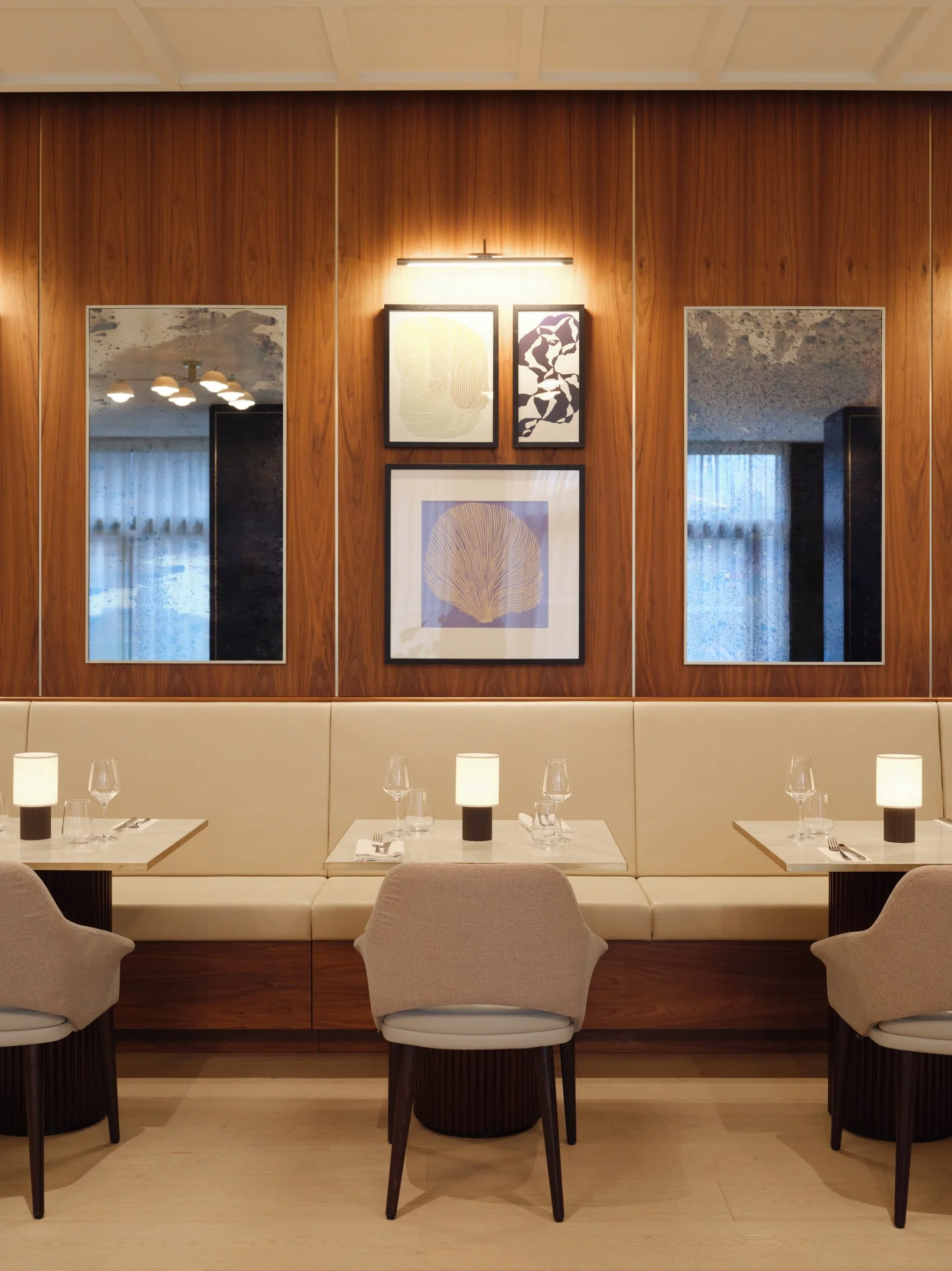 A modern restaurant interior with a beige upholstered bench, three beige chairs, and three tables set with glasses, napkins, and lamps. The wood-paneled wall has three framed art pieces and a window with curtains reflecting in two mirrors.