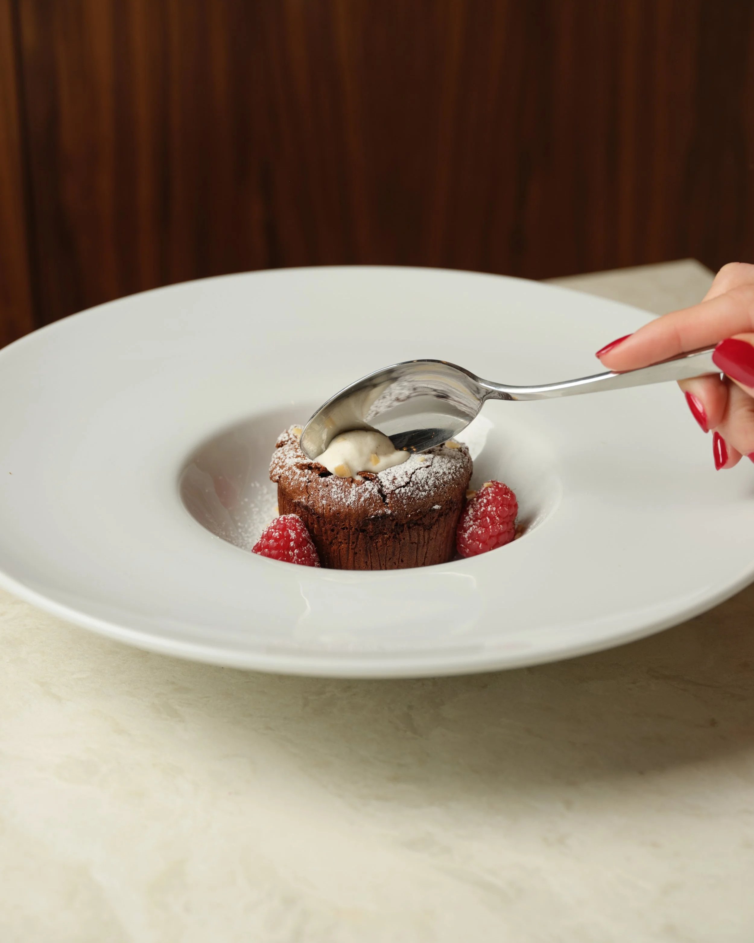 A chocolate lava cake on a white plate, garnished with powdered sugar and strawberries, with a hand holding a spoon about to scoop it.