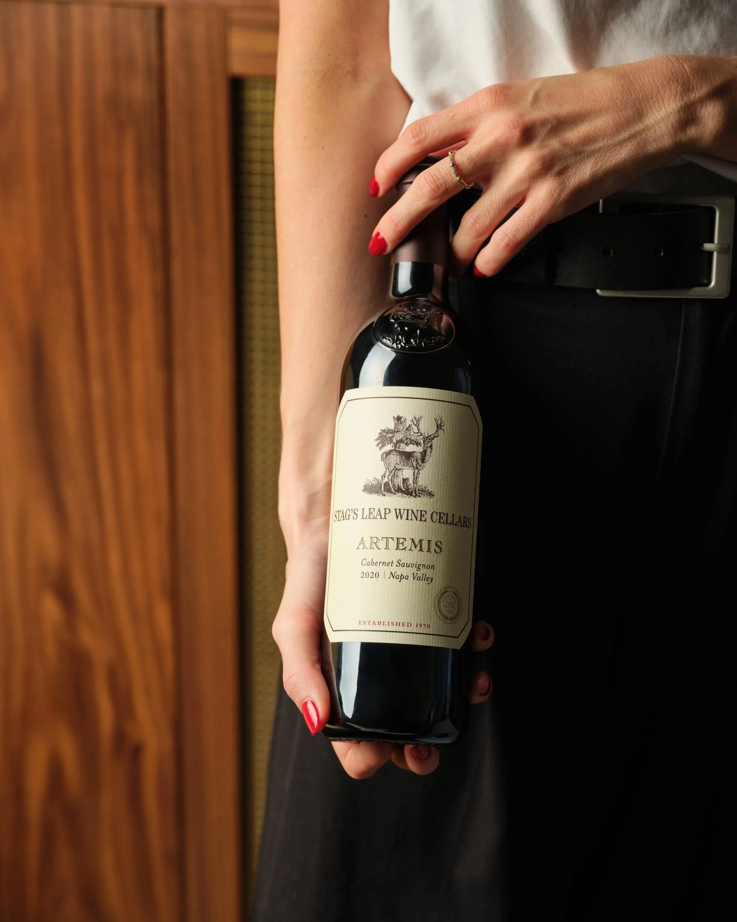 A person's hand with red painted nails holding a bottle of Artemis Cabernet Sauvignon wine from Stag's Leap Wine Cellars.