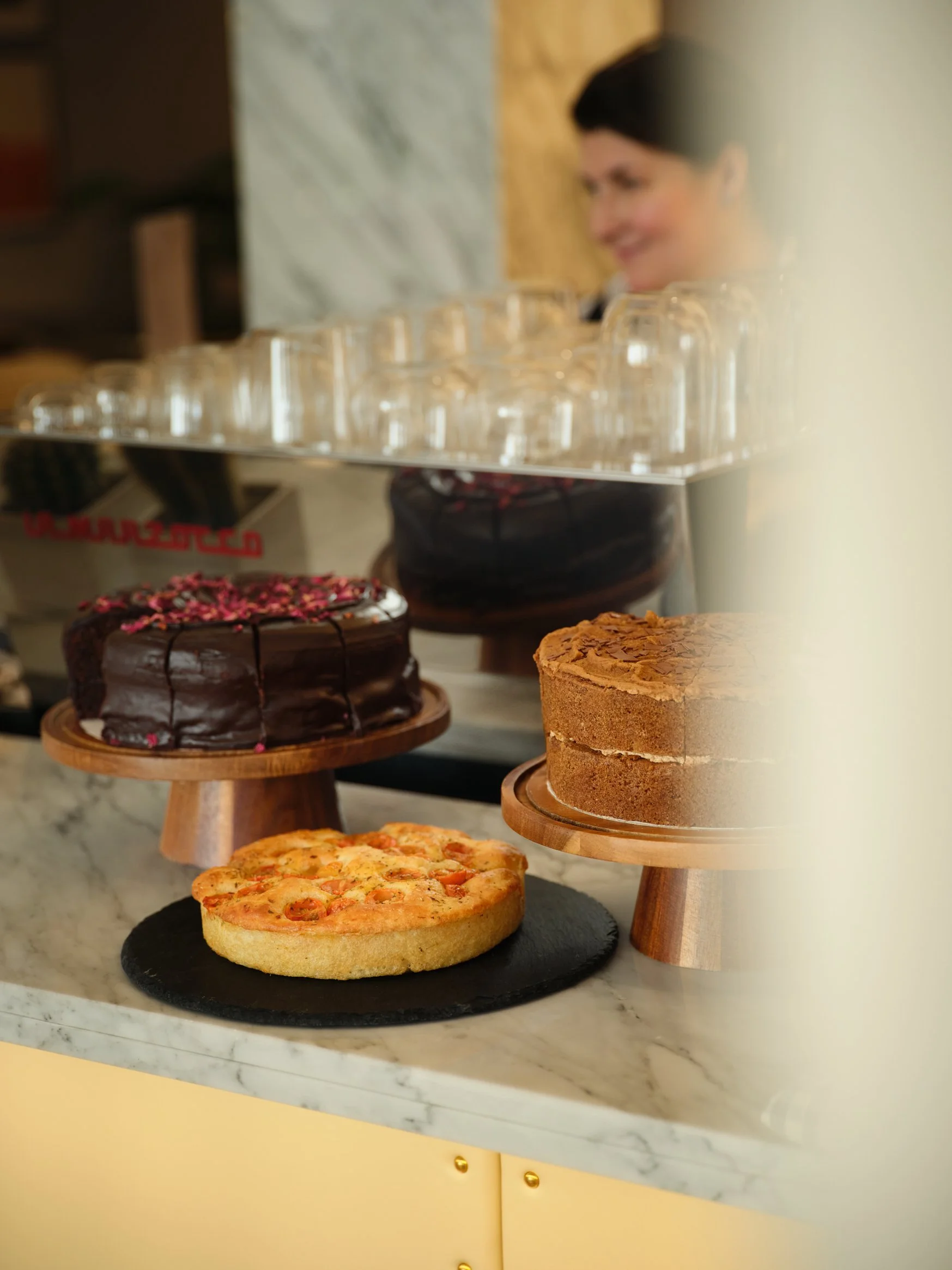 Assortment of cakes on display counter with a woman in the background.