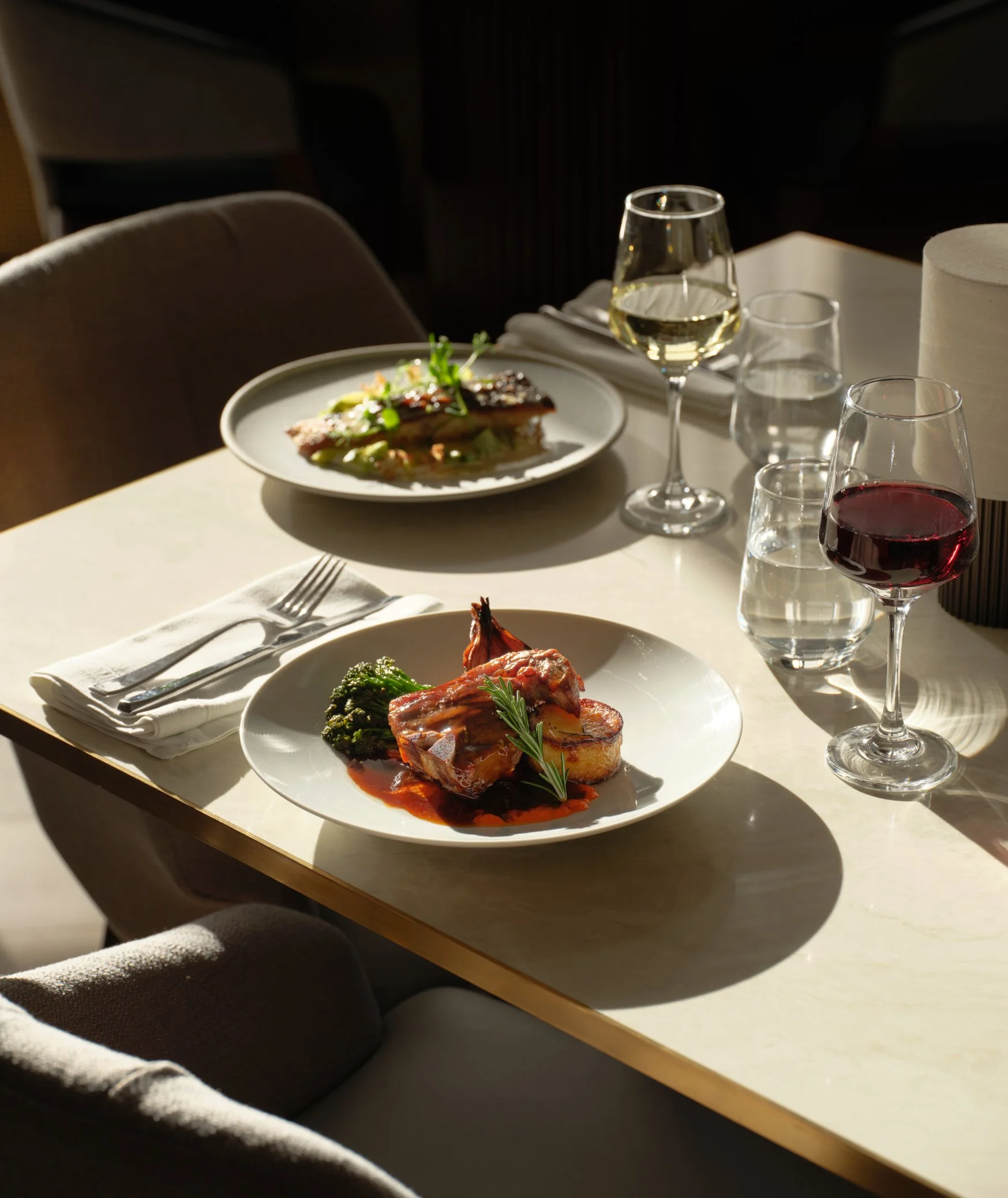 Elegant dining table set with two plates of gourmet food, two glasses of red wine, a glass of white wine, and water glasses, illuminated by natural light.