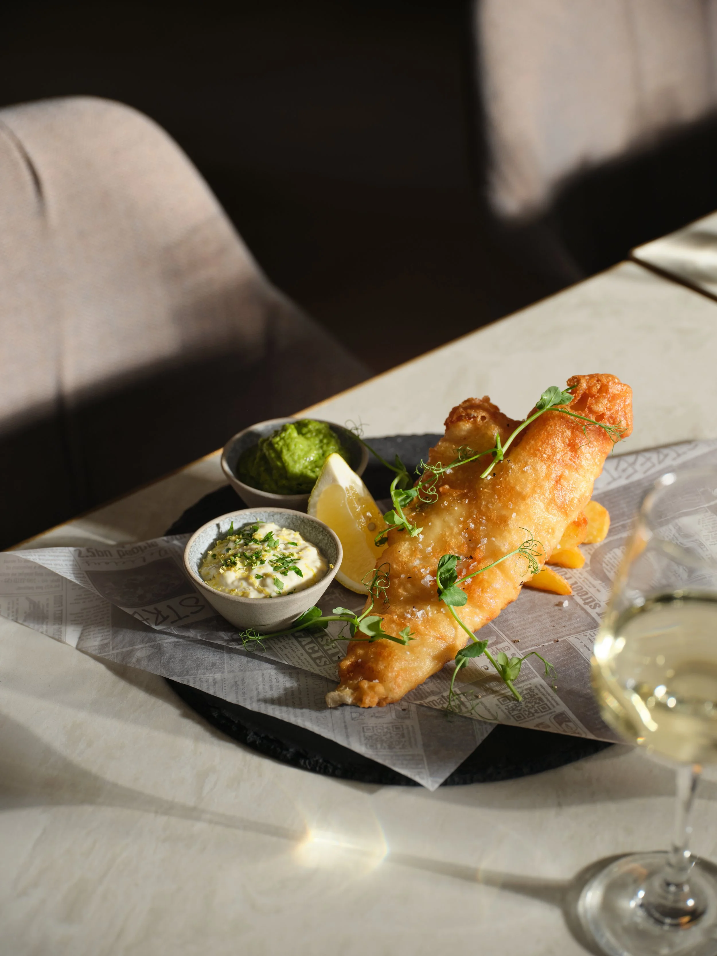 Fried fish filet served with lemon, green sauce, and tartar sauce on a black plate with newspaper lining, accompanied by a glass of white wine.
