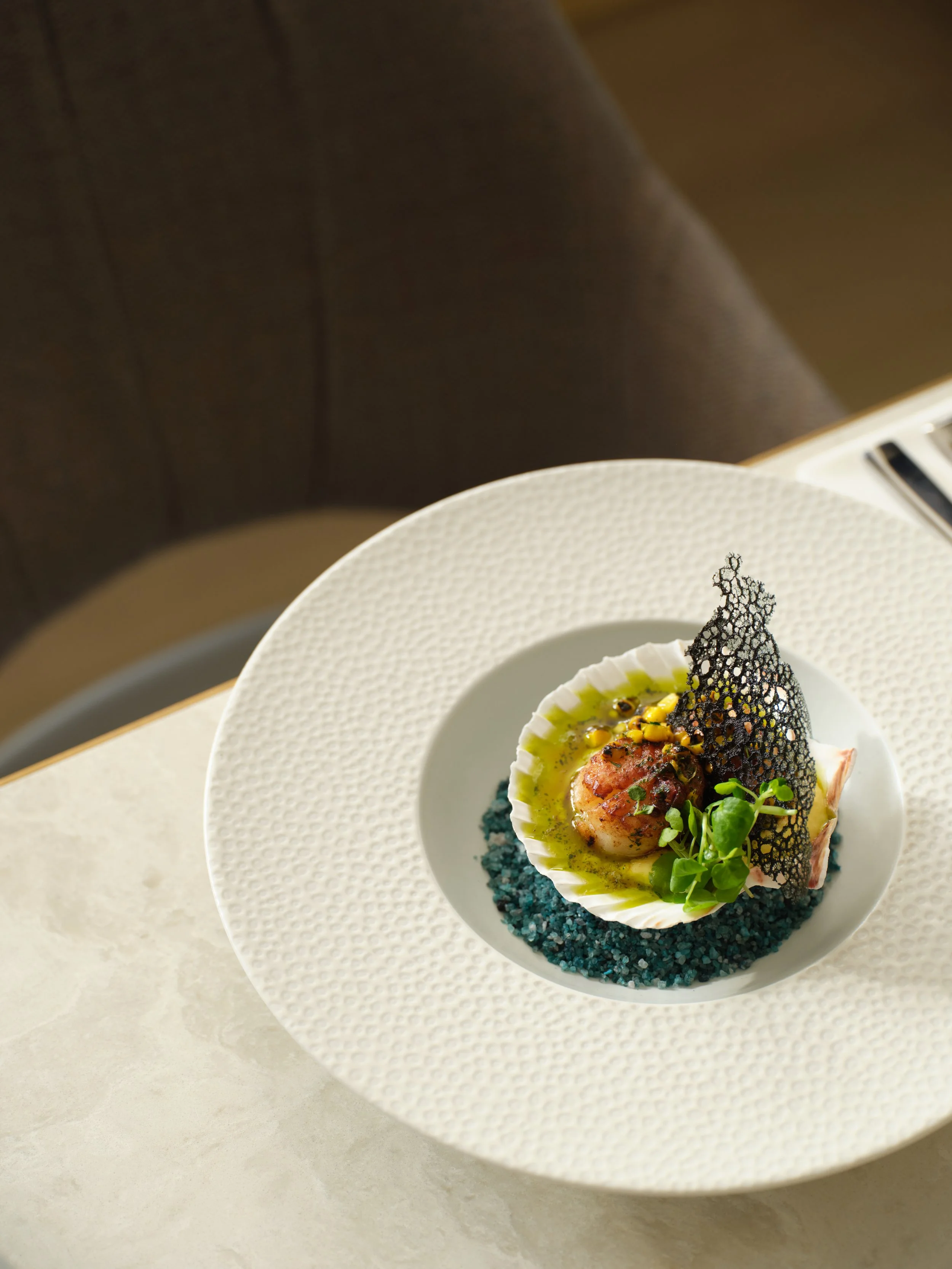 A gourmet dish with a scallop, green sauce, microgreens, a black lace-like garnish, and blue salt on a white textured plate.