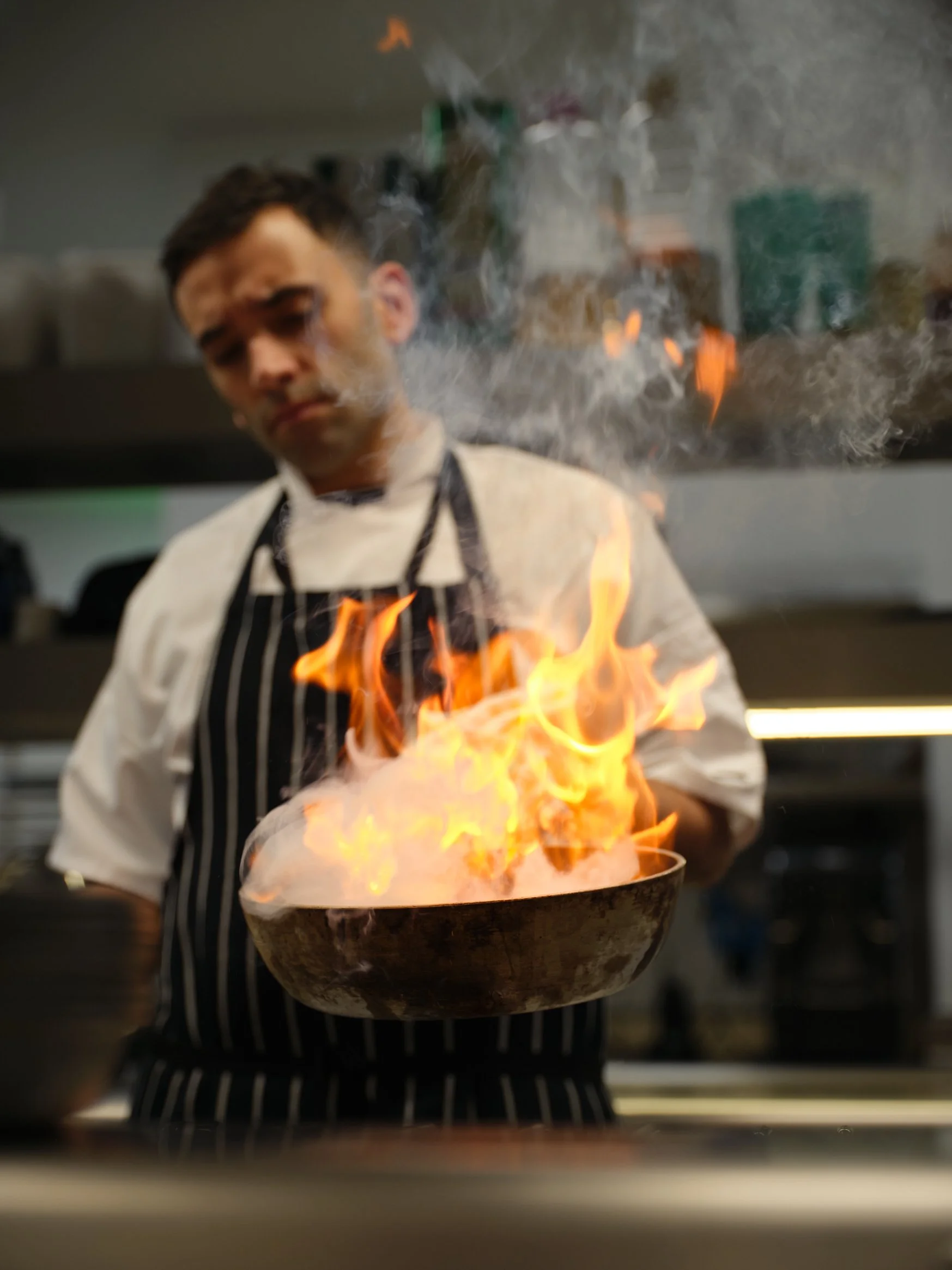 Chef holding a frying pan with open flames in a professional kitchen.