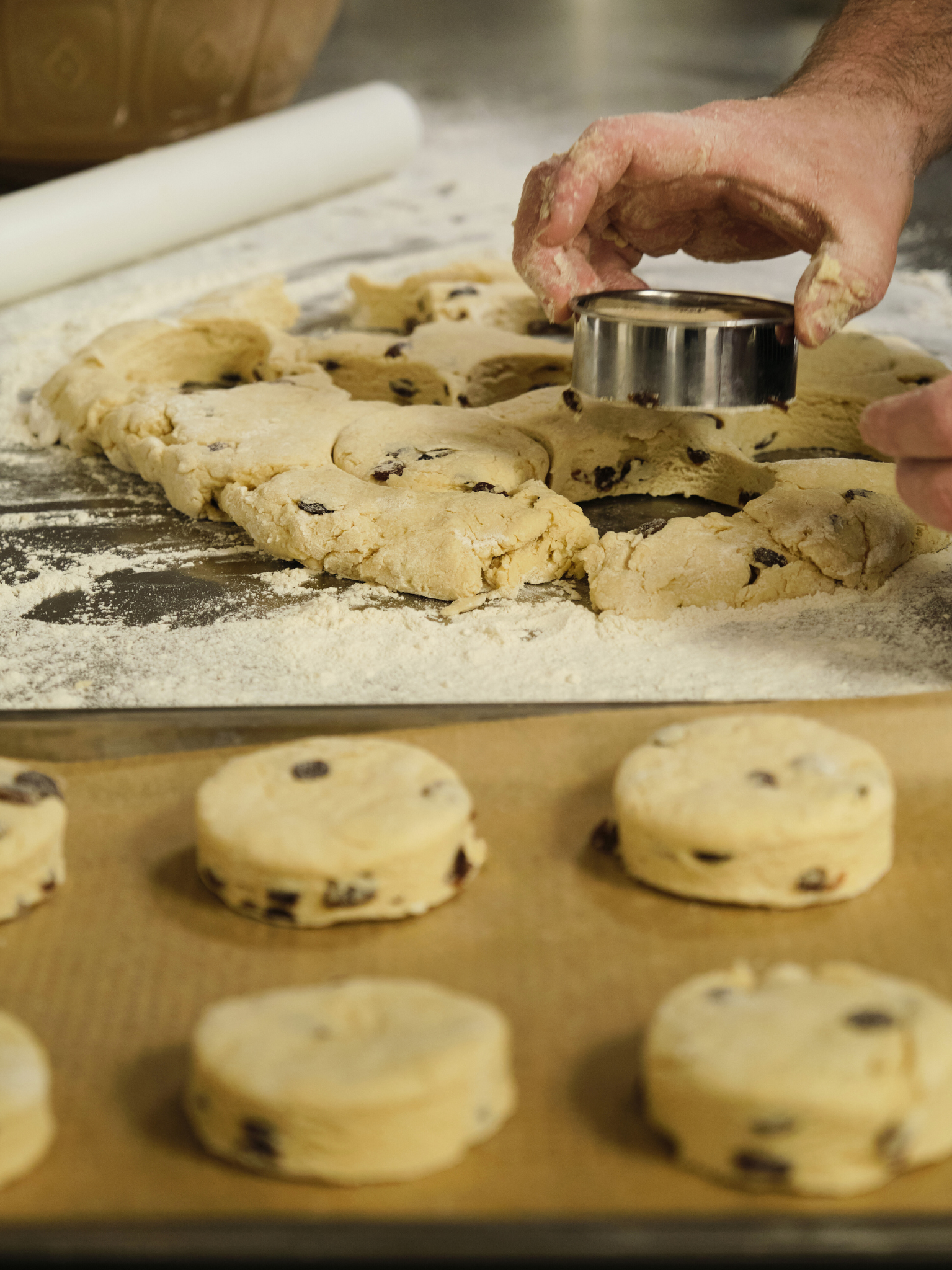 Baker cutting fruit scones with a round cutter on a floured surface, with scone cutouts on a baking sheet.