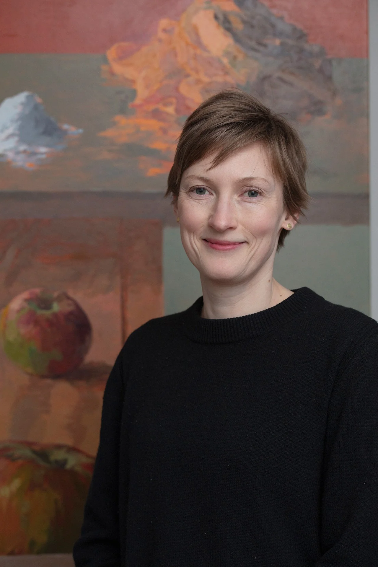 A woman with short brown hair wearing a black sweater, standing in front of a colorful abstract painting of mountains, clouds, and a fruit.