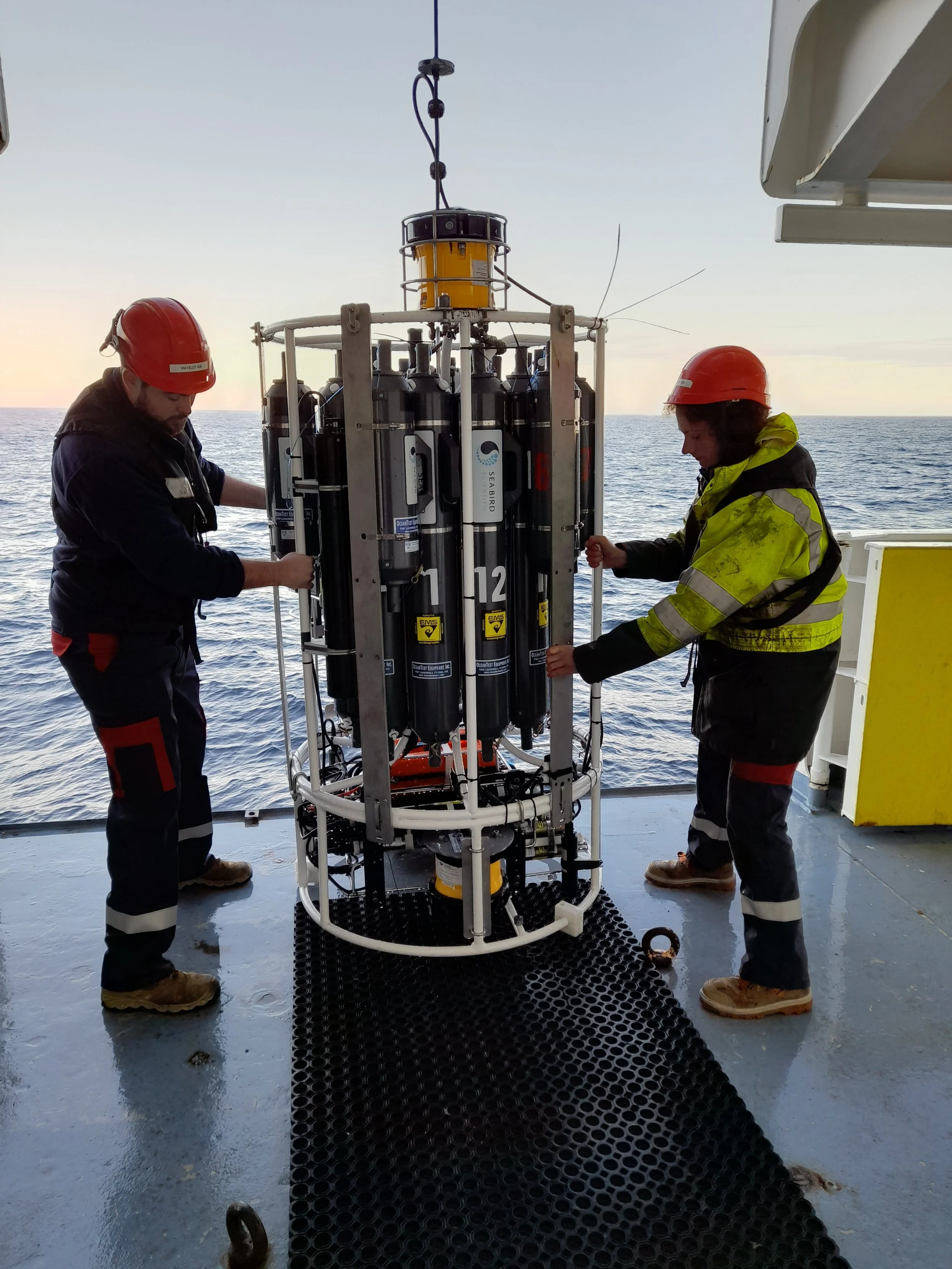 Two workers in safety gear installing or maintaining a piece of scientific equipment on a ship at sea during sunset.