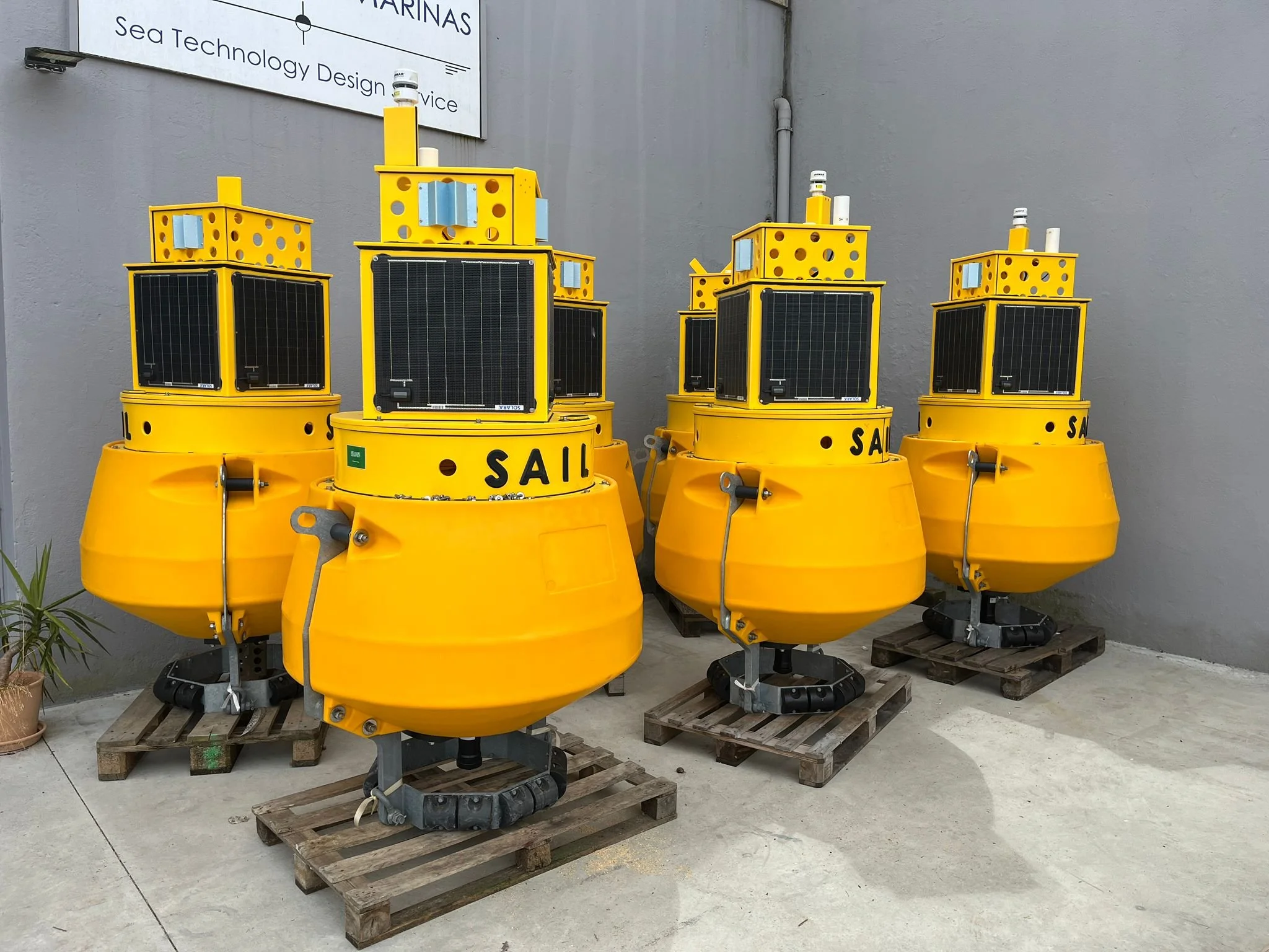 Four yellow autonomous surface vehicles with solar panels, stacked on wooden pallets against a gray wall.