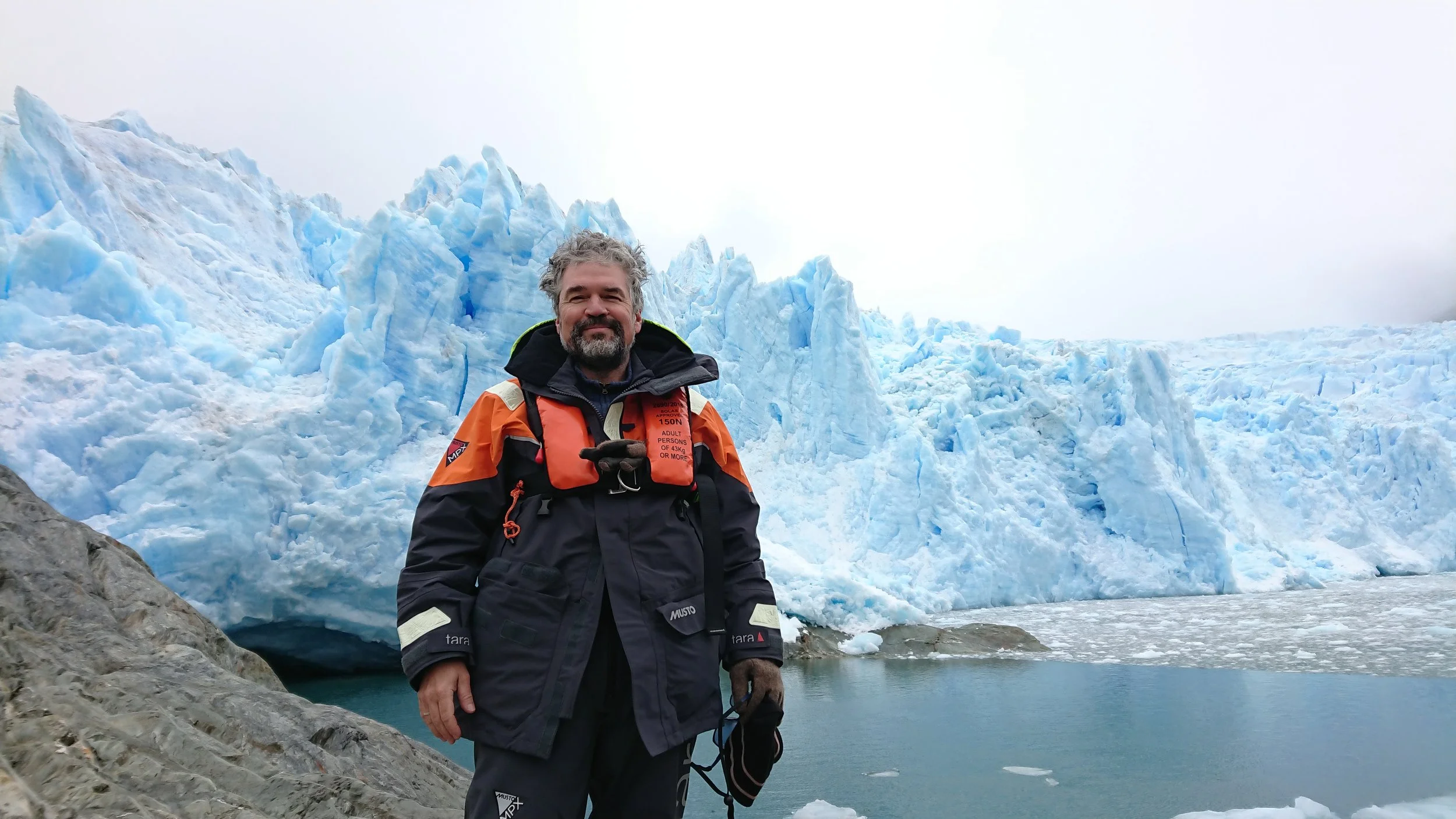 Man in outdoor gear standing in front of a glacier and icy water.