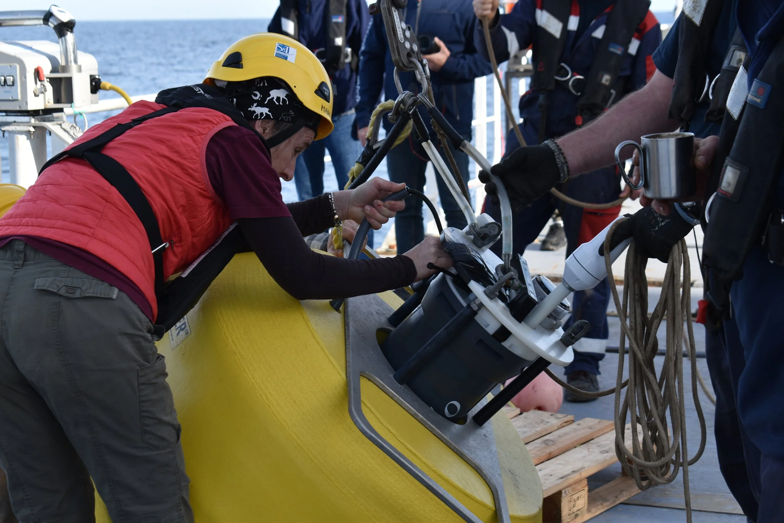 Scientists and crew aboard a research vessel preparing equipment for an oceanographic experiment, with the ocean visible in the background.